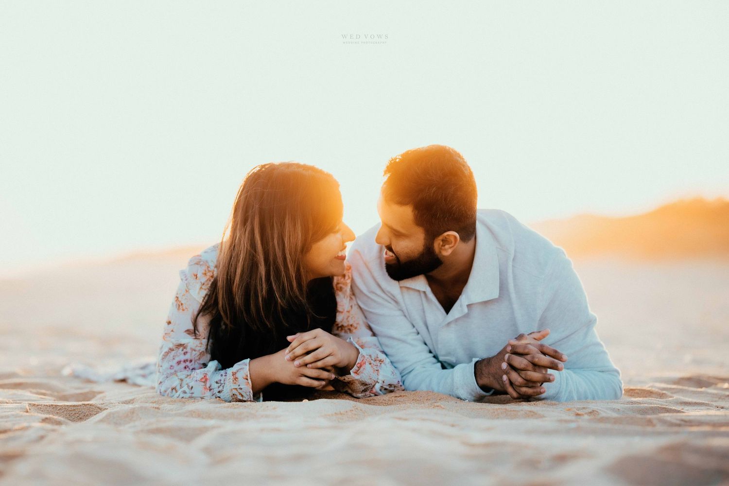 Couple lying on golden sand beach face to face, sharing an intimate romantic moment at sunset.