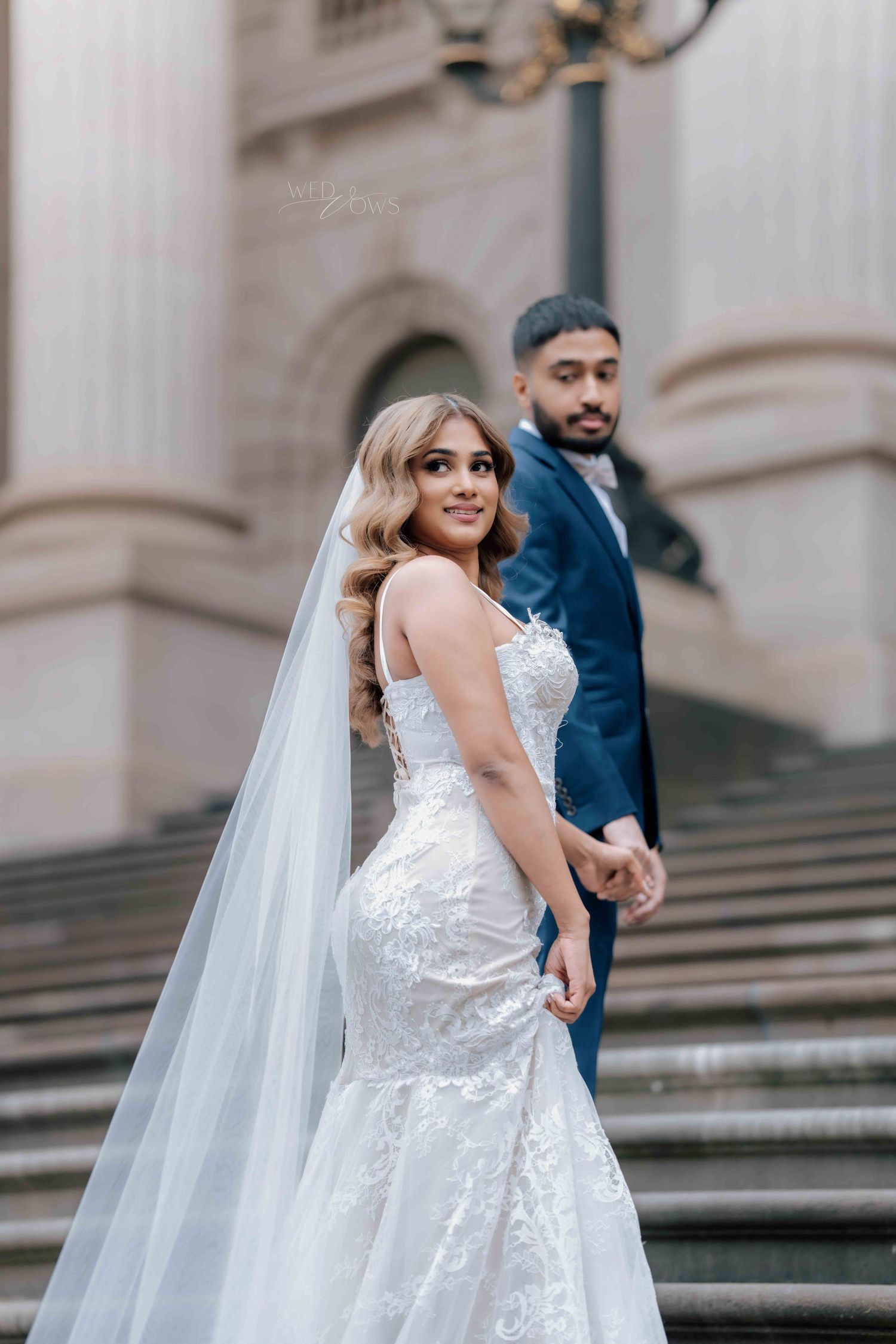 Smiling bride in lace mermaid gown with long veil leading groom down grand stone steps.