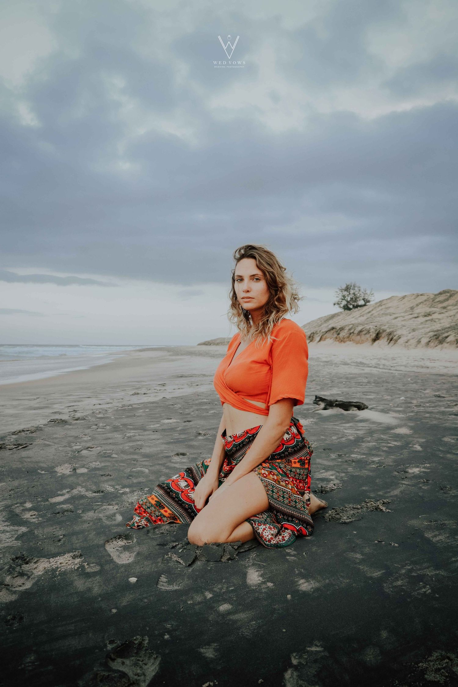 Woman in orange crop top and patterned skirt kneeling on a dark sandy beach under cloudy sky.