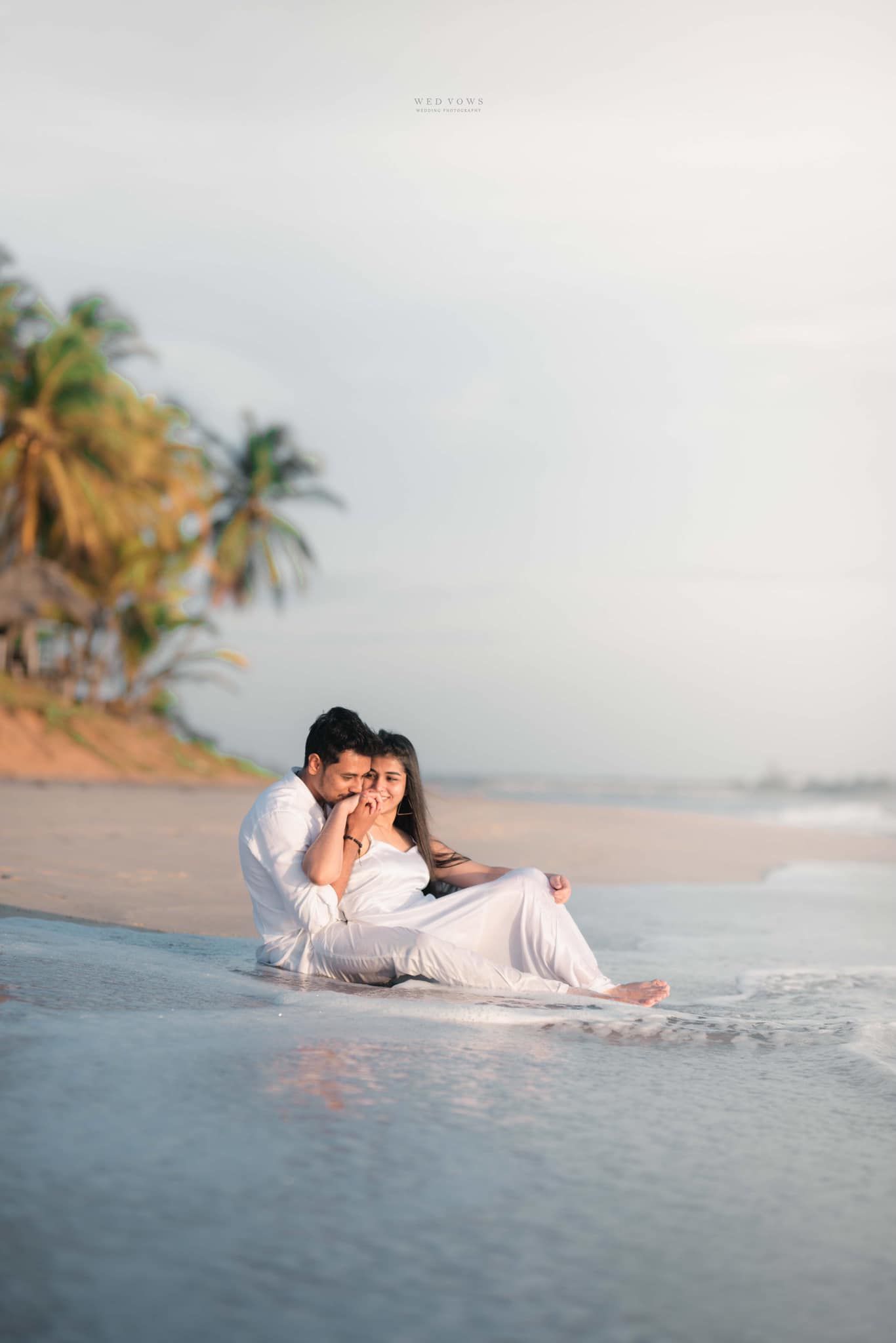 Romantic couple sitting together in white outfits on a tropical beach shore with gentle waves and palm trees.