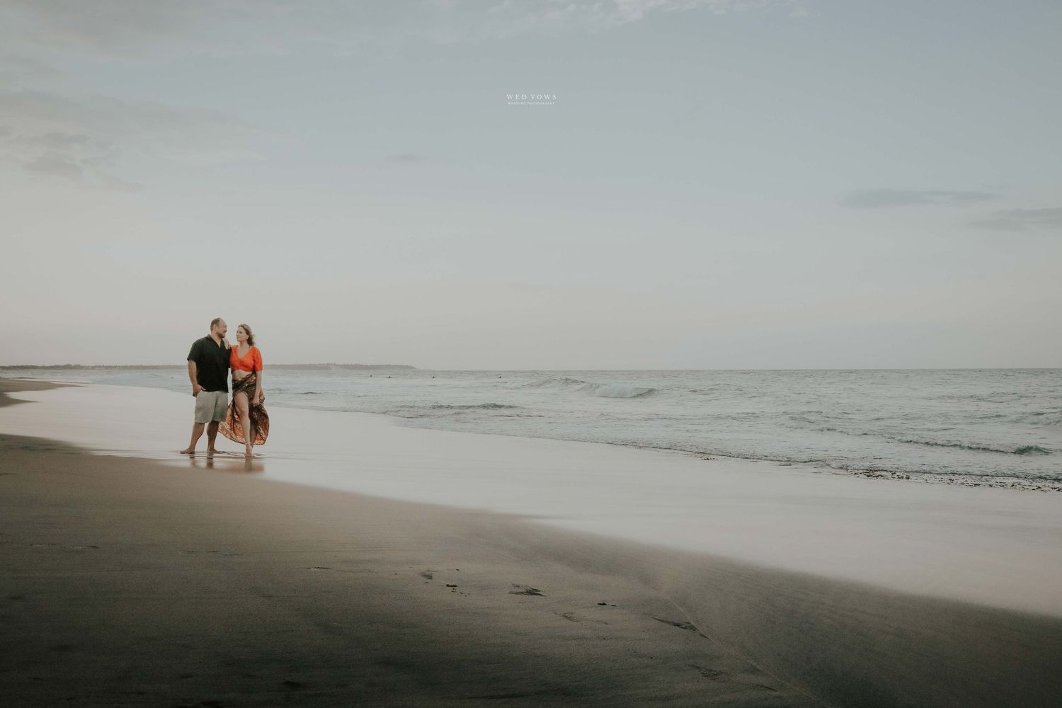 Couple walking along shoreline with a dog on a calm beach at sunset, holding hands.