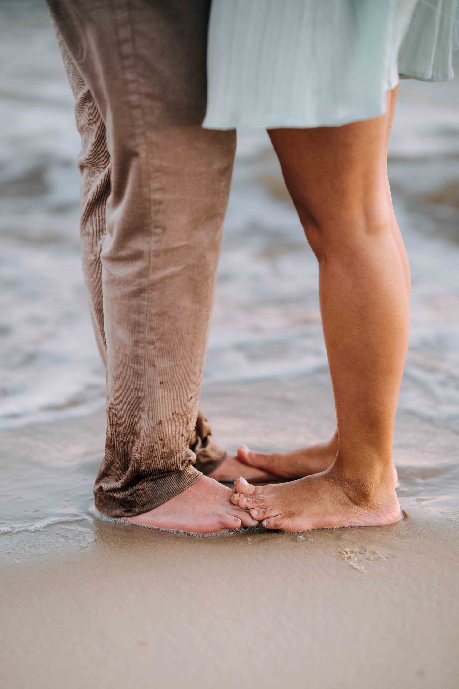 Close-up of couple's bare feet touching on wet sandy beach, woman in blue dress, man in tan pants.