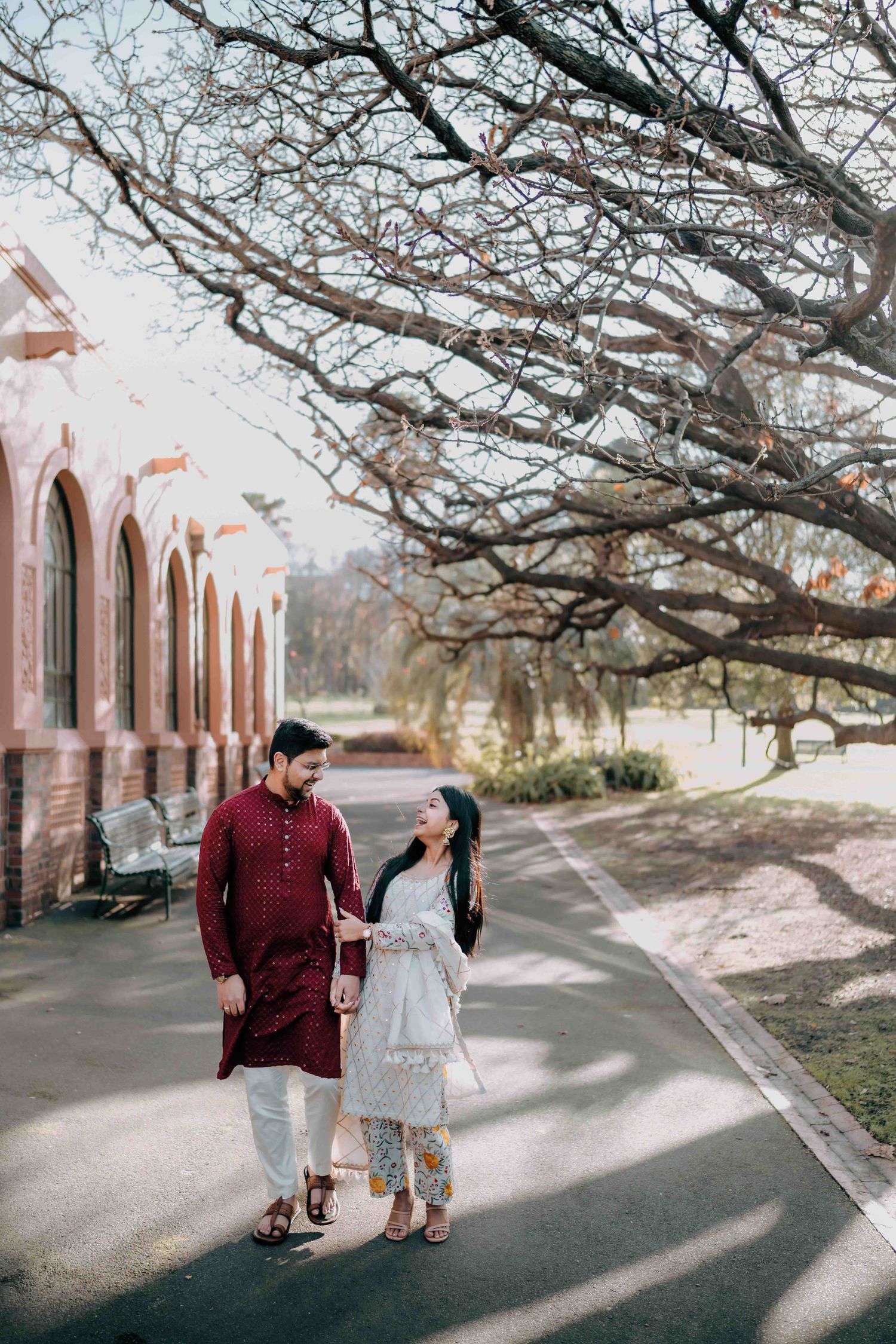 South Asian couple in kurta outfits walking and smiling near ornate red brick building outdoors.