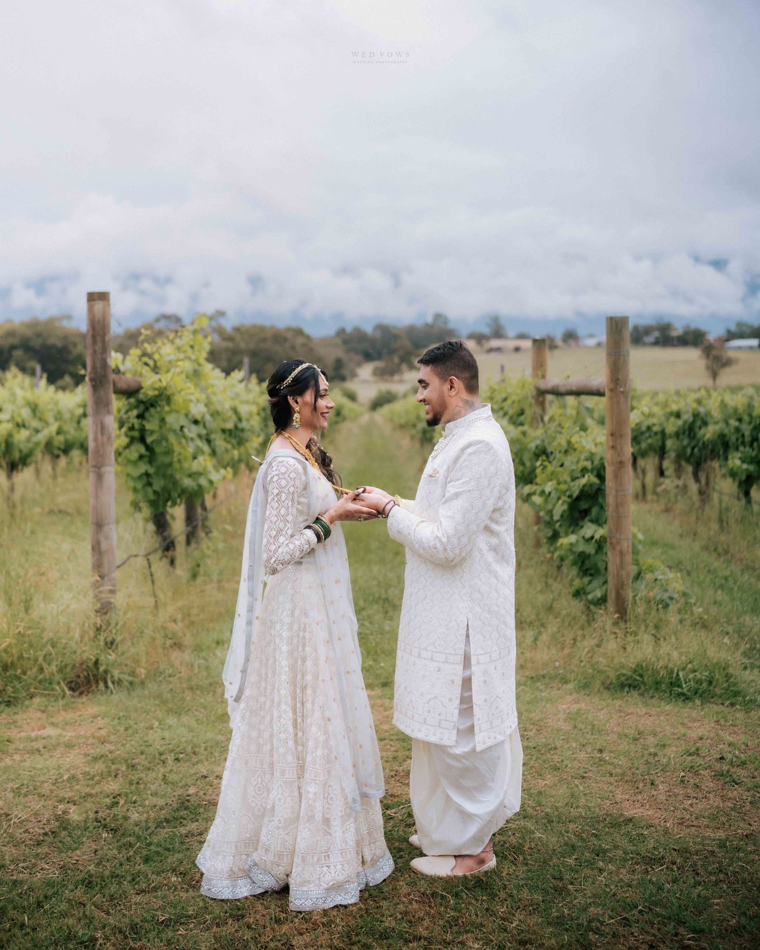 Indian couple in white wedding attire exchange vows in a lush vineyard setting.