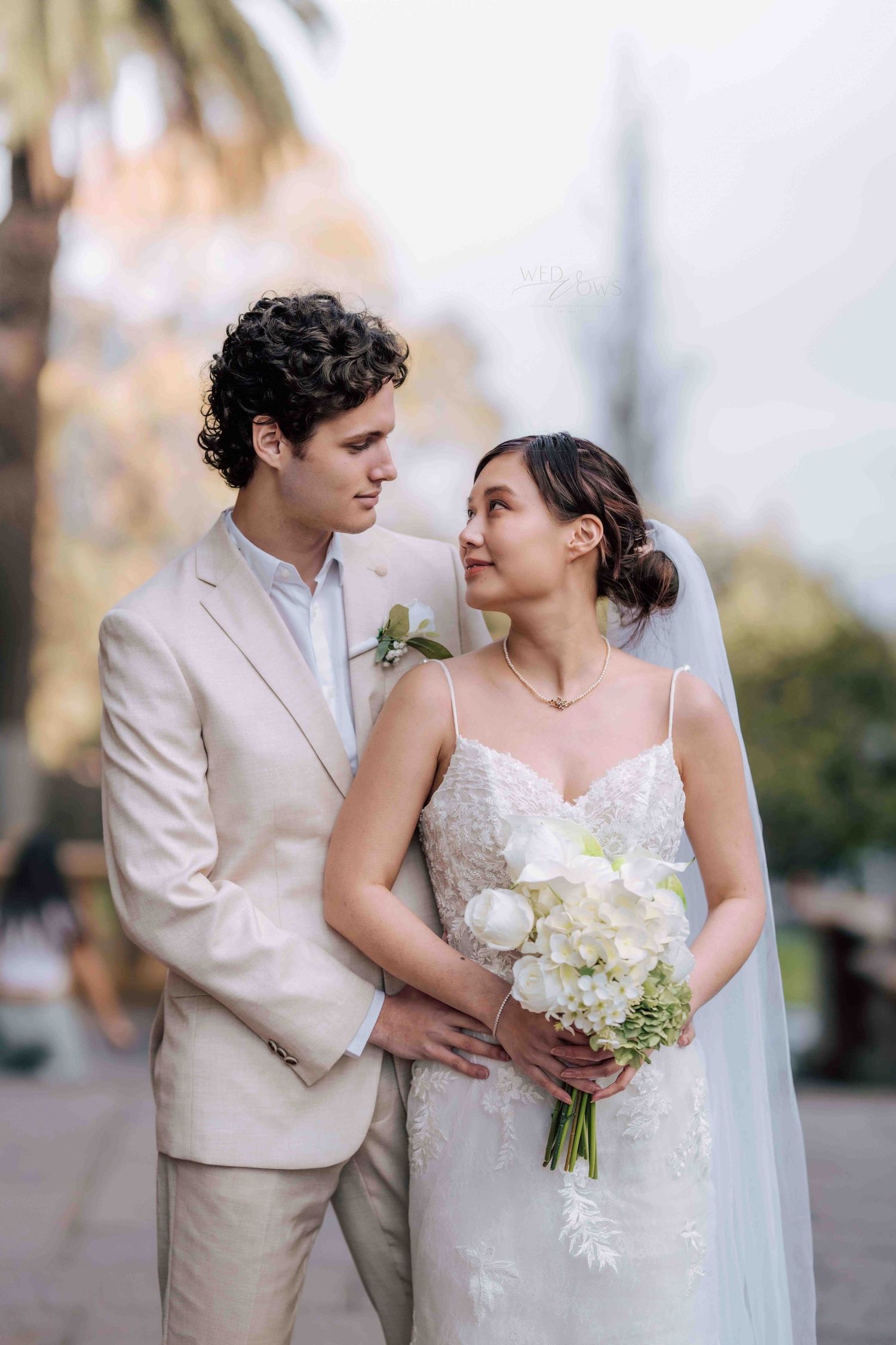 Groom in beige suit gazes lovingly at bride holding white bouquet in lace gown and veil in outdoor setting.