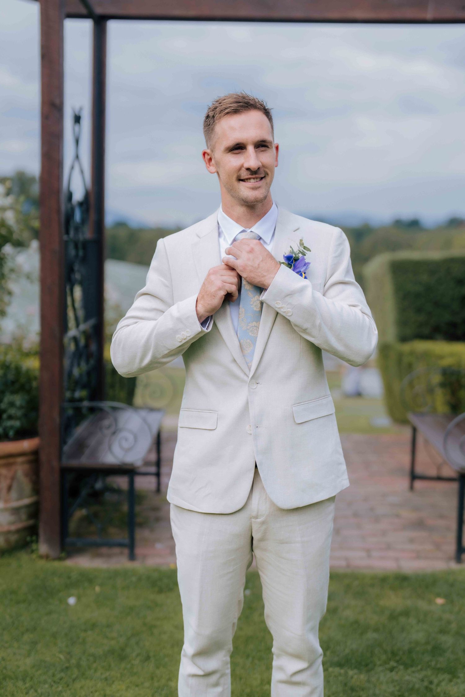 Smiling groom in ivory suit adjusts tie with blue floral boutonniere at outdoor wedding.