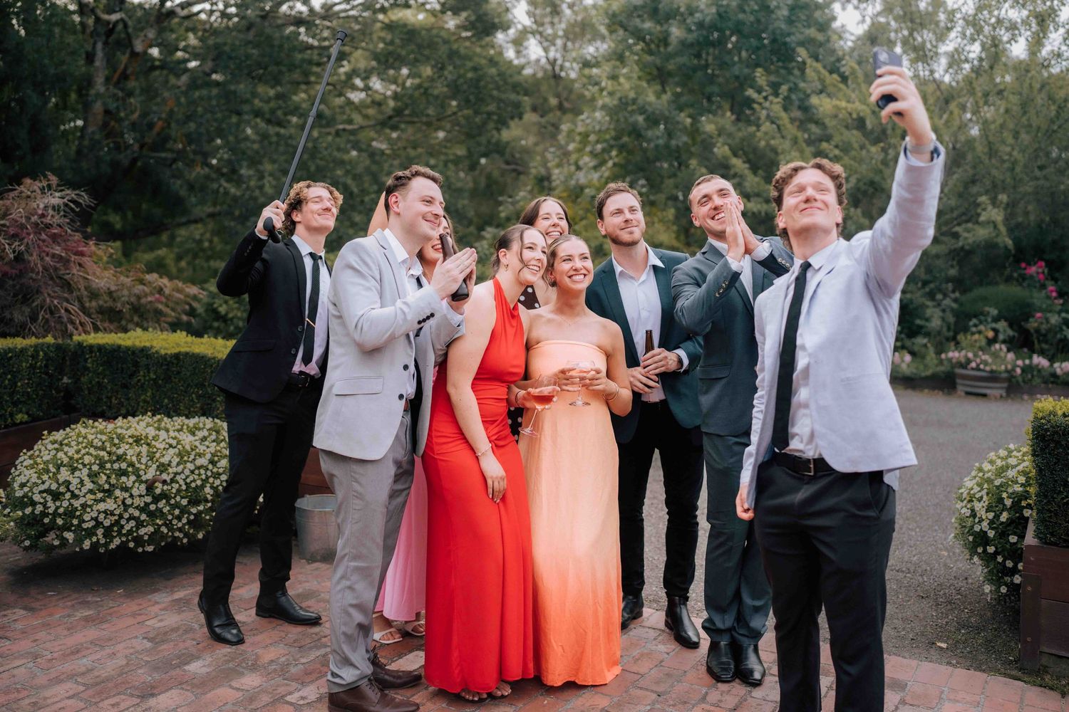Wedding guests in formal attire take a group selfie with a selfie stick in a garden.