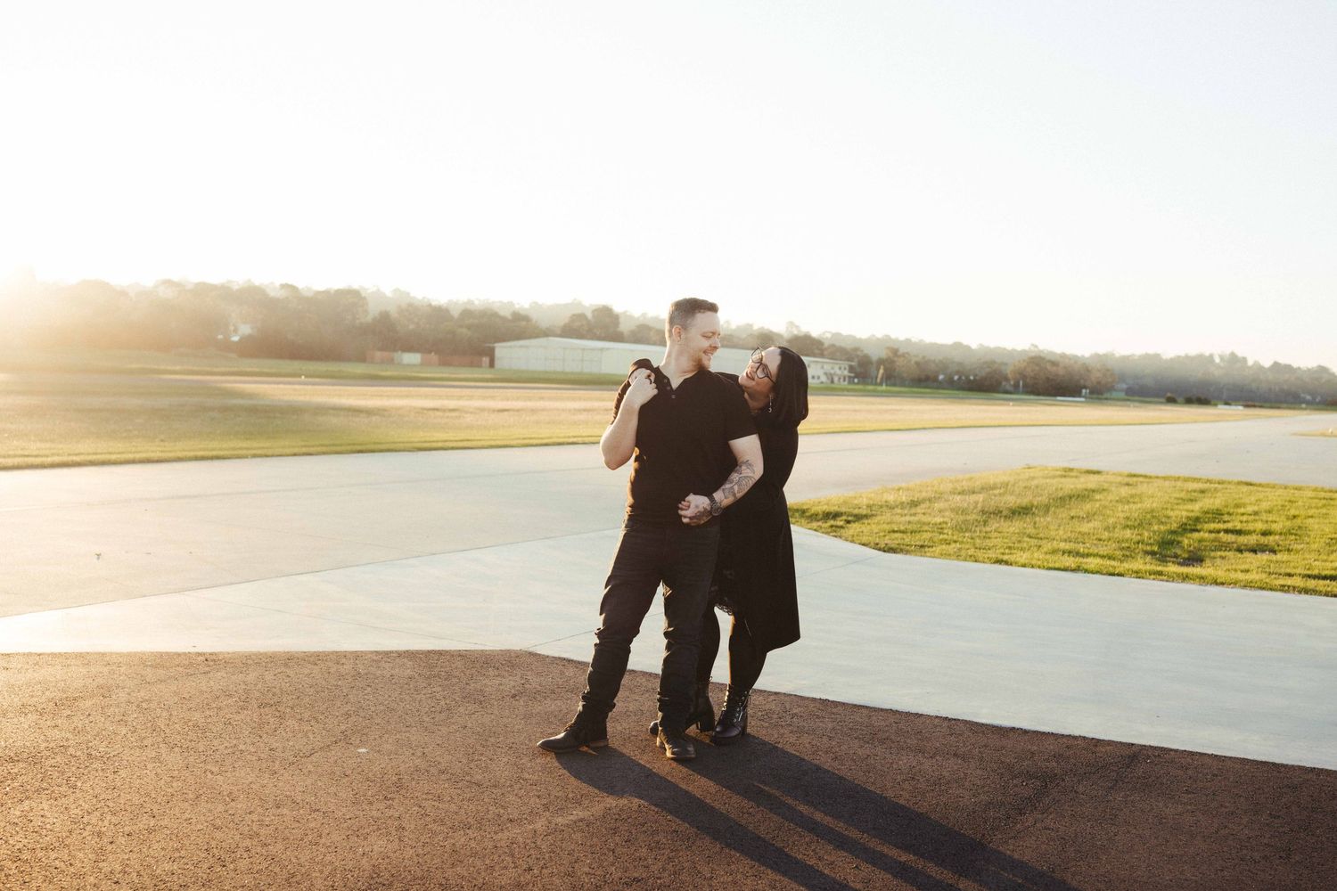 Couple sharing a romantic kiss at sunset on an airport tarmac, dressed in black outfits.