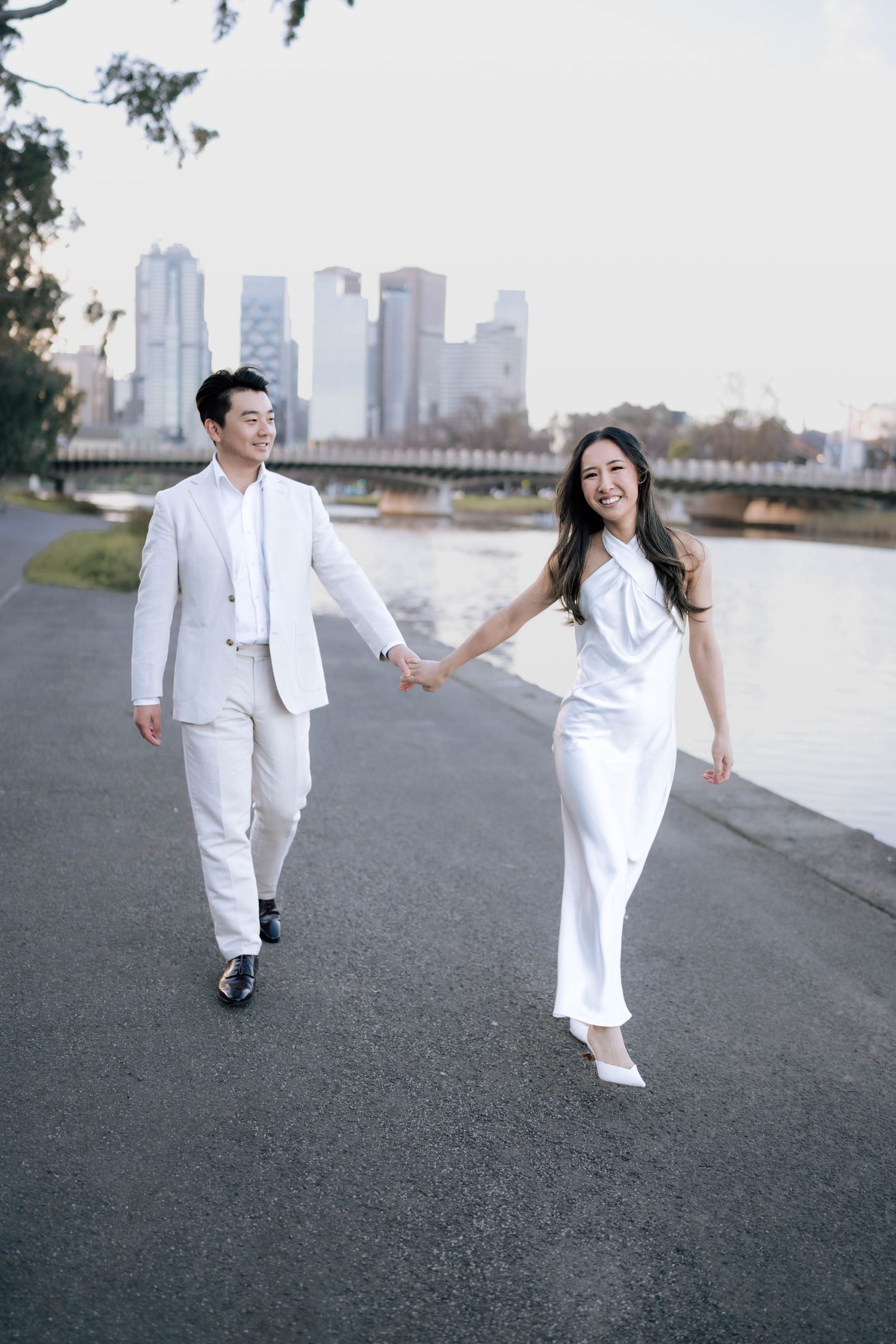 Couple in white outfits holding hands and walking along a riverside path with city skyline in background.