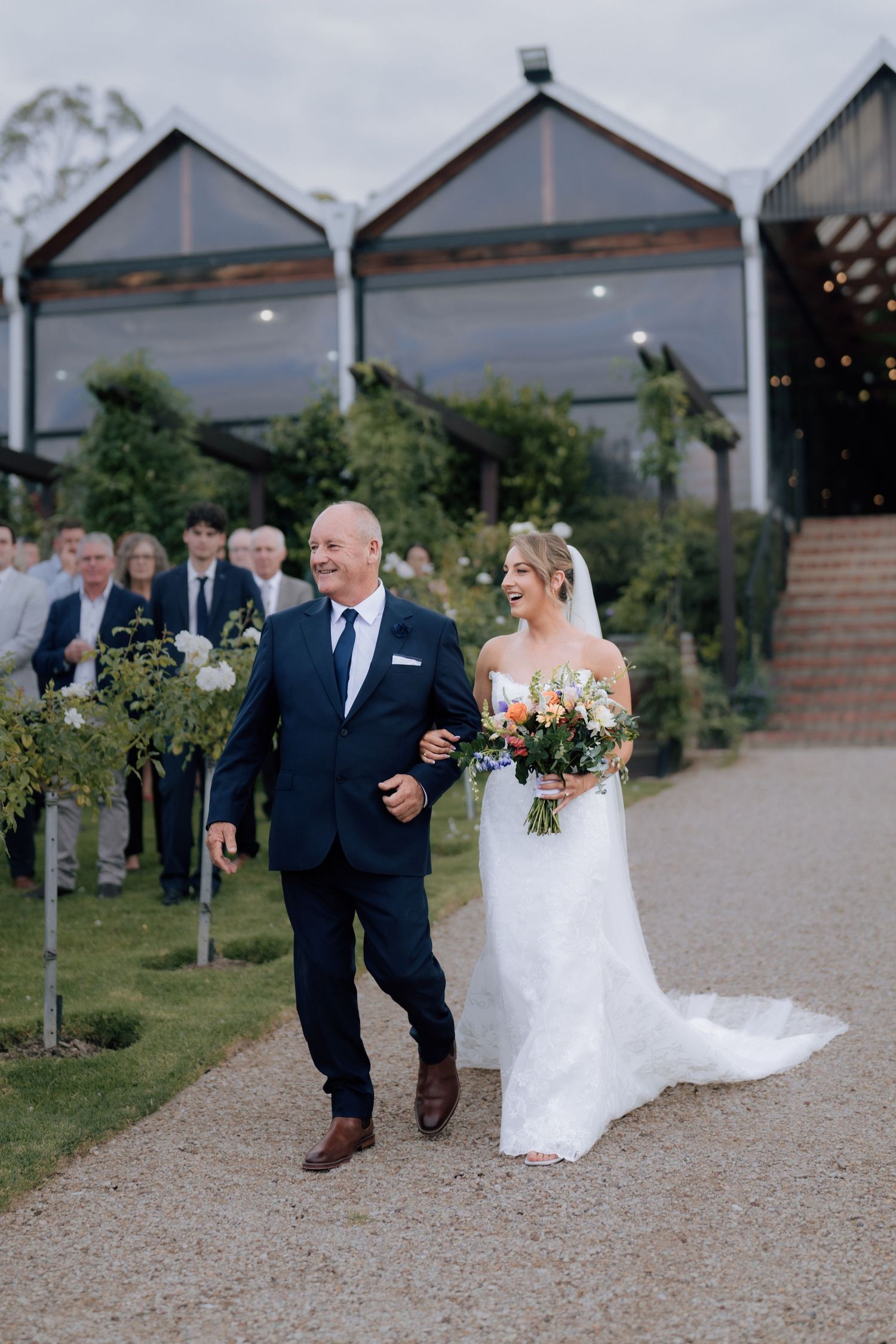 Bride in strapless white gown walking down aisle with father at outdoor vineyard wedding.