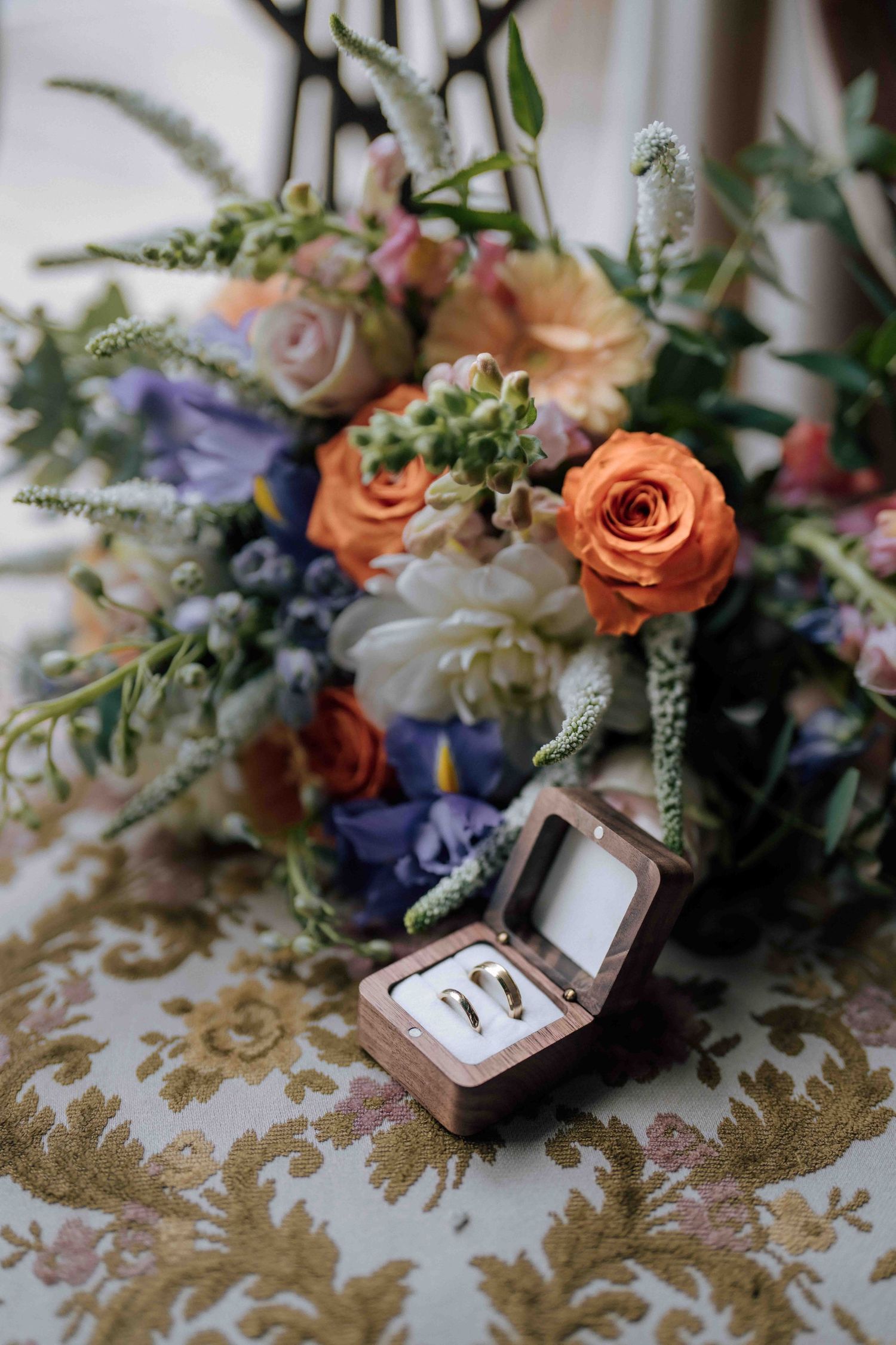 Gold wedding rings in a wooden box beside a vibrant bridal bouquet of orange, purple, and white flowers.