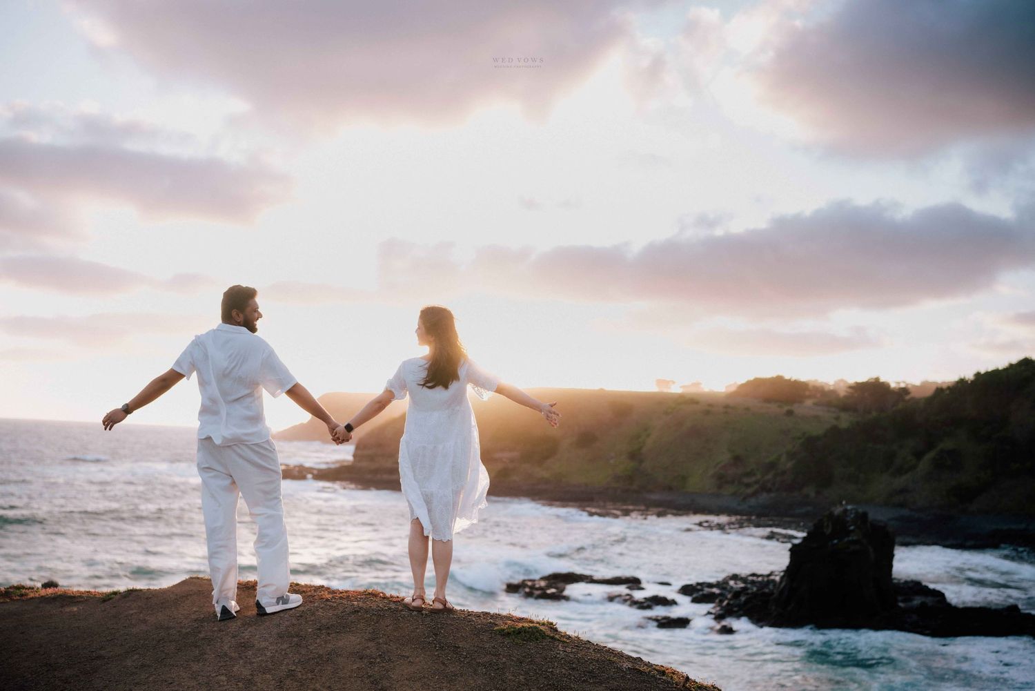 Couple in white holding hands on coastal cliff at sunset with ocean waves and rocky shoreline.