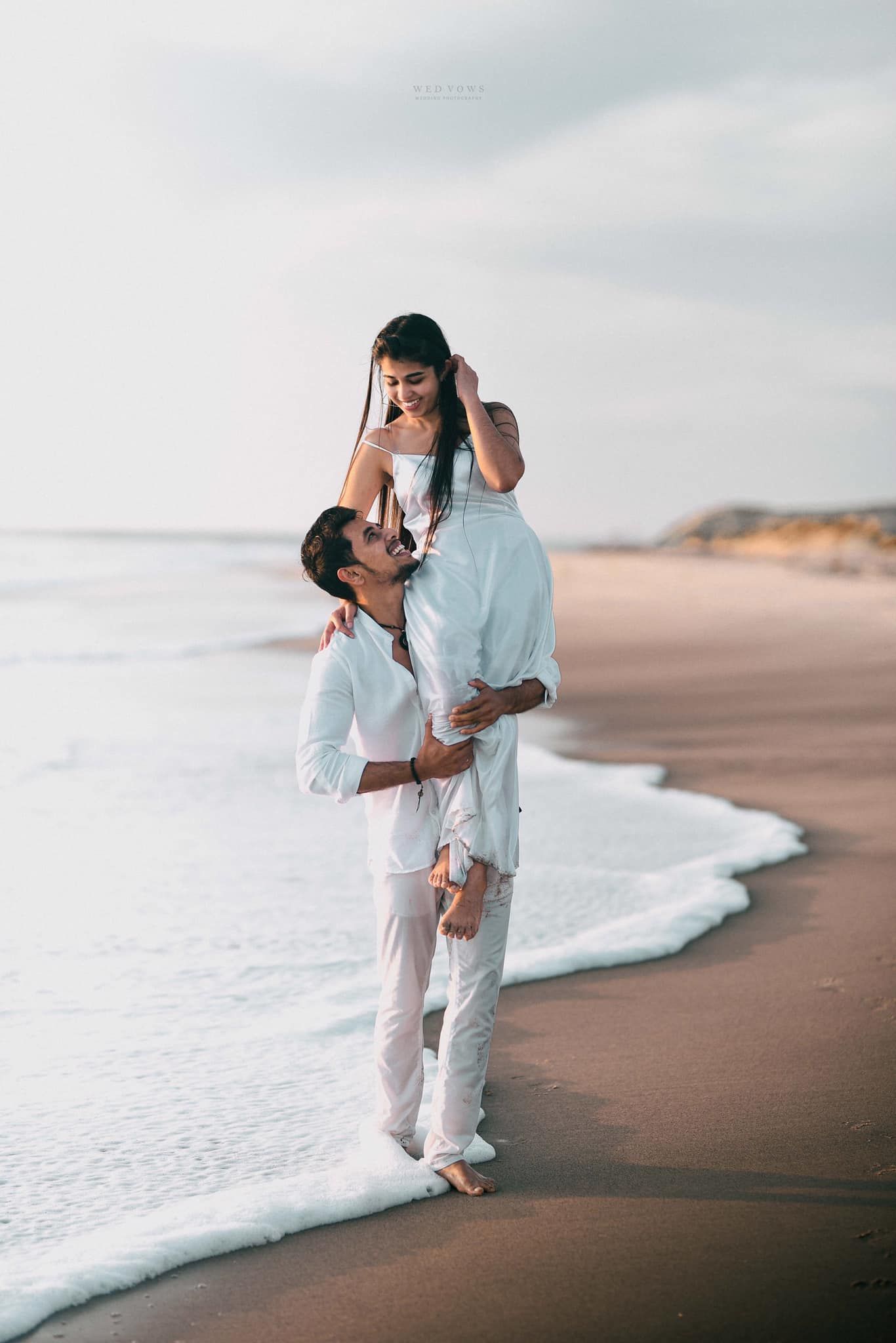 Man in white outfit lifting woman on a sandy beach with ocean waves at their feet.