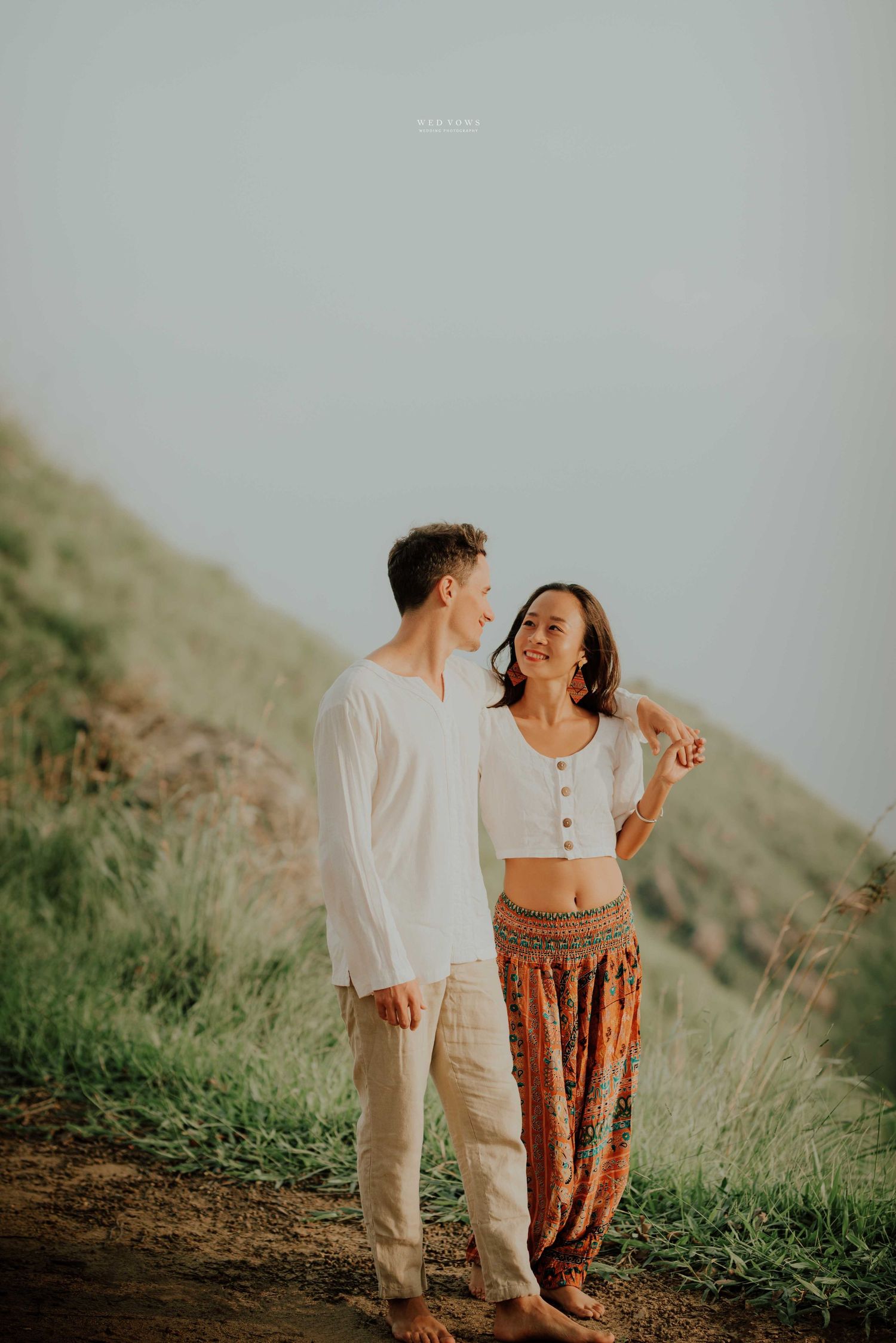 A smiling couple in bohemian outfits walks together on a grassy hillside, woman in crop top and tribal print harem pants.