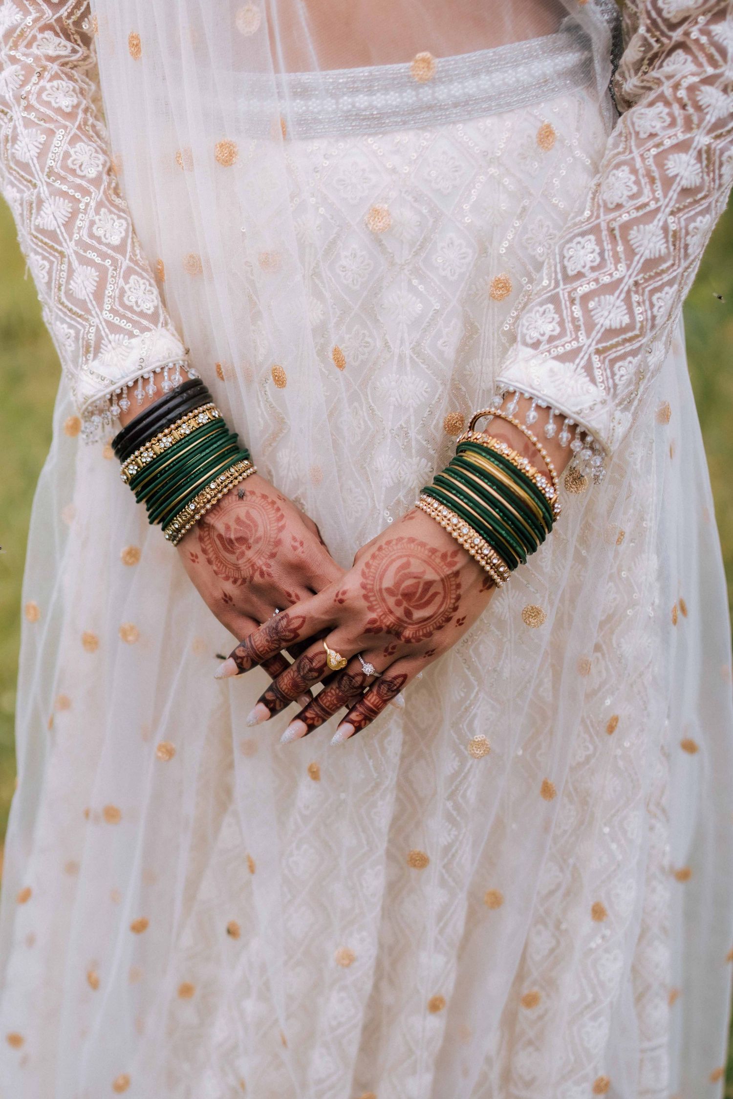 Close-up of Indian bride's hands with intricate henna mehndi and green and gold bangles on white lehenga.