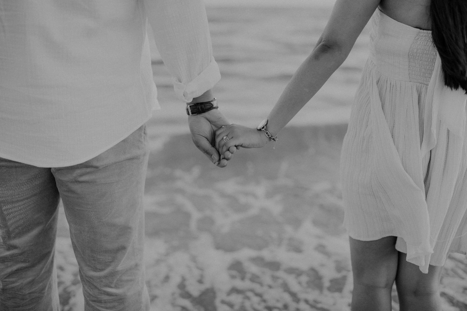 Close-up black and white photo of couple holding hands on a beach with ocean backdrop.