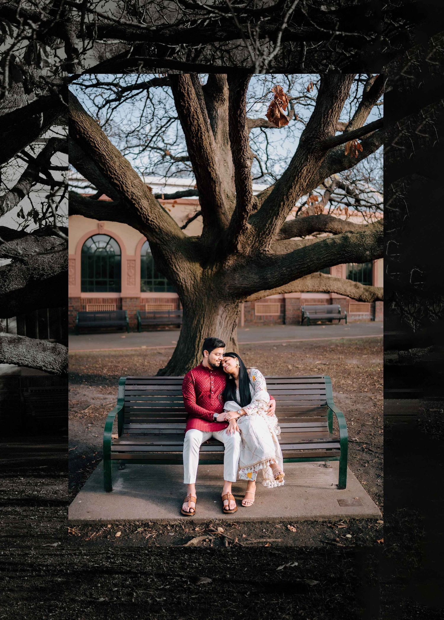 Couple in traditional attire sitting closely on a park bench beneath a large bare tree in winter.