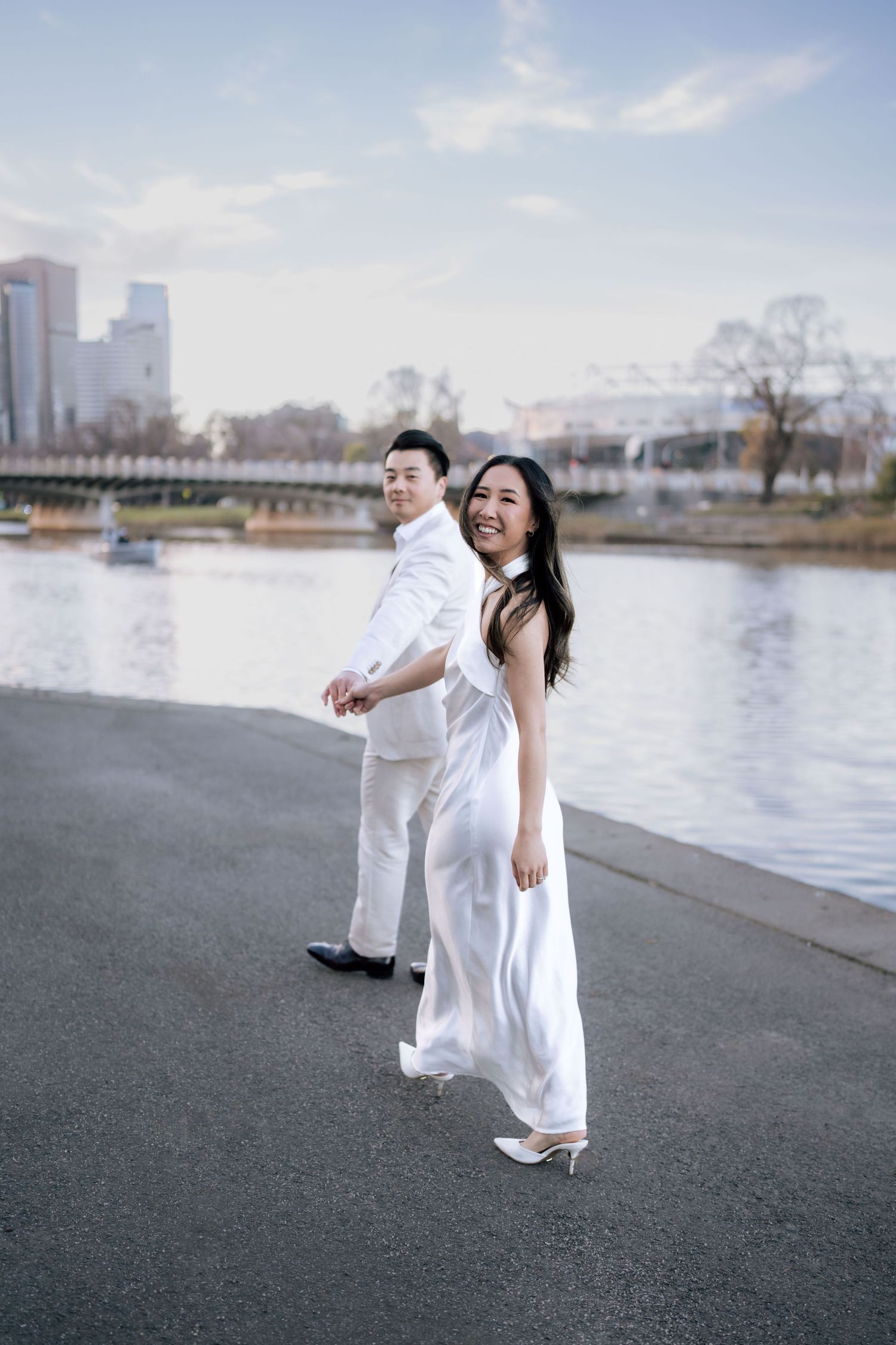 Smiling couple in white outfits walking hand in hand along a riverside path with city skyline.