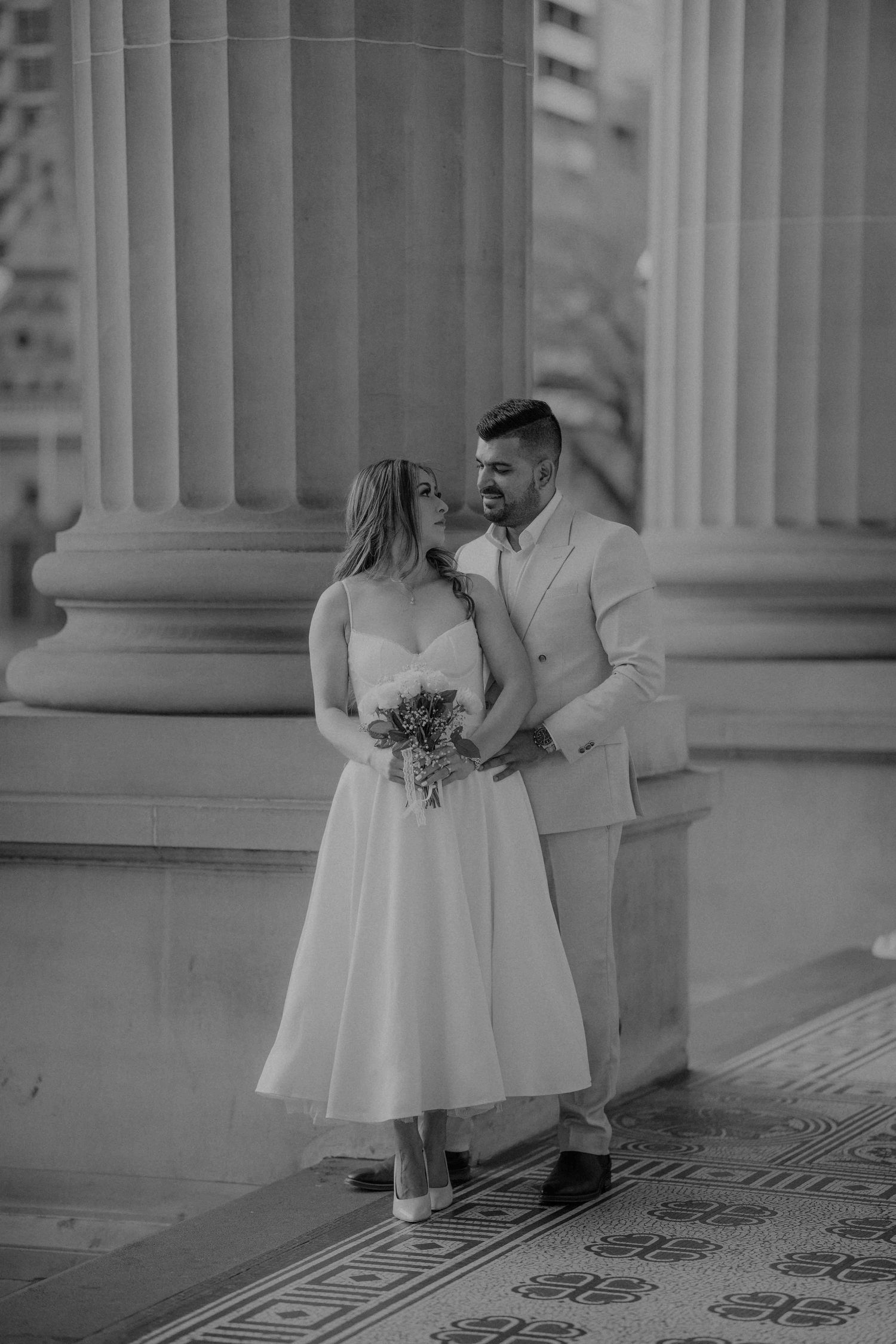 Black and white portrait of a loving couple embracing beside tall stone columns at a historic venue.