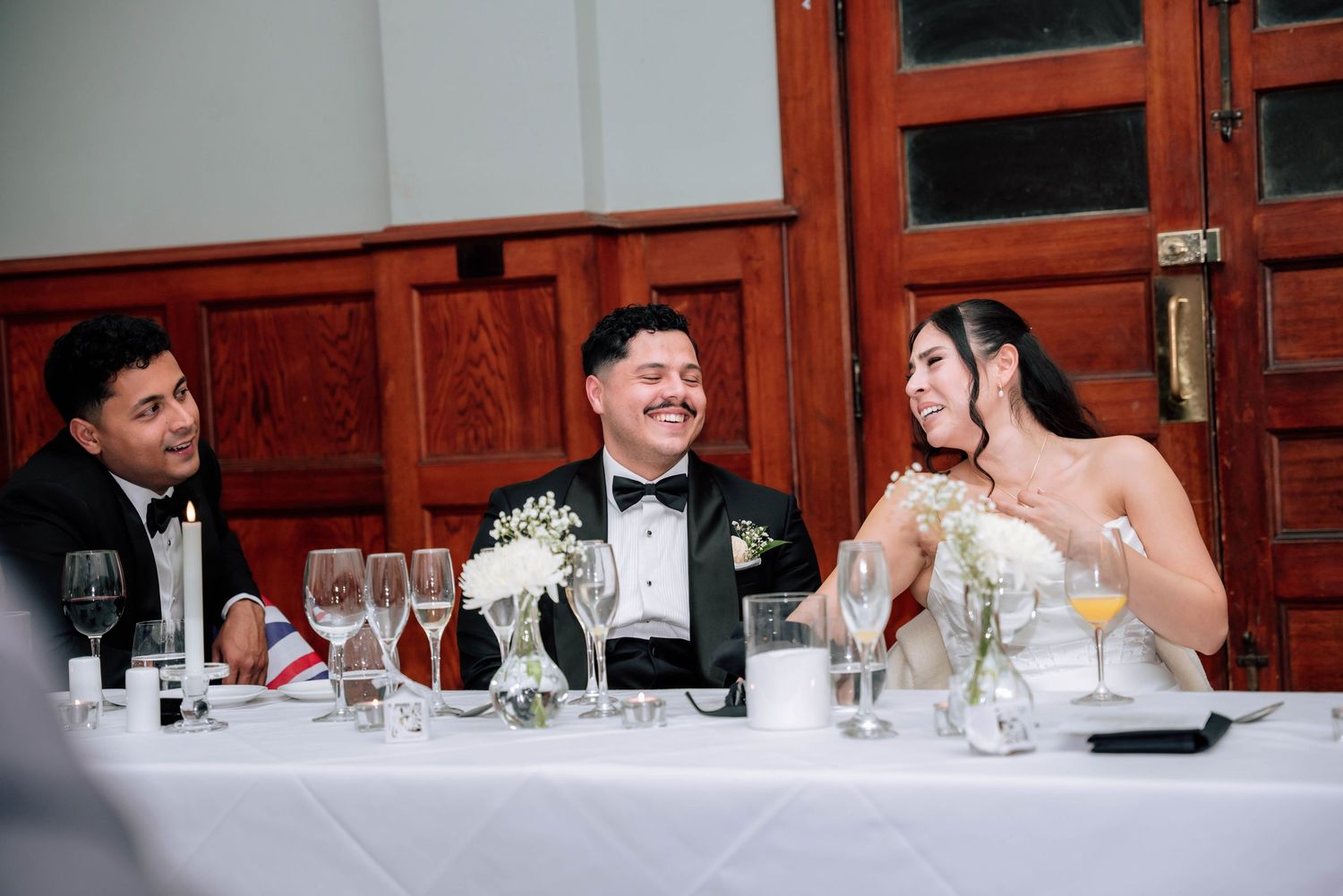 Bride and groom laugh with guest at elegant wedding reception head table with white floral decor.