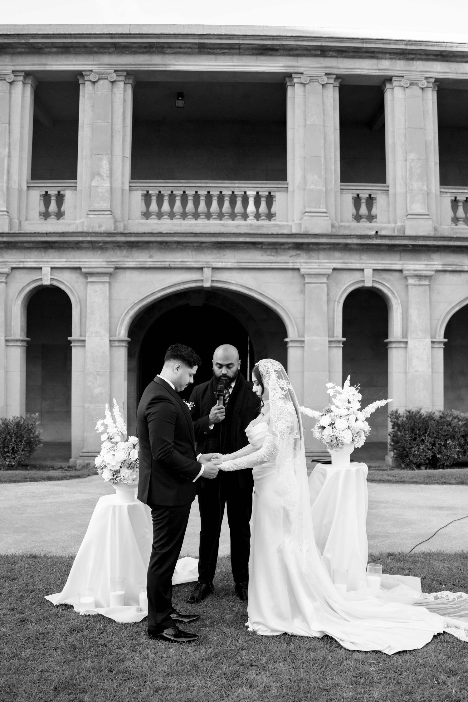 Black and white photo of a bride and groom exchanging vows outdoors before an elegant stone building with arched columns.