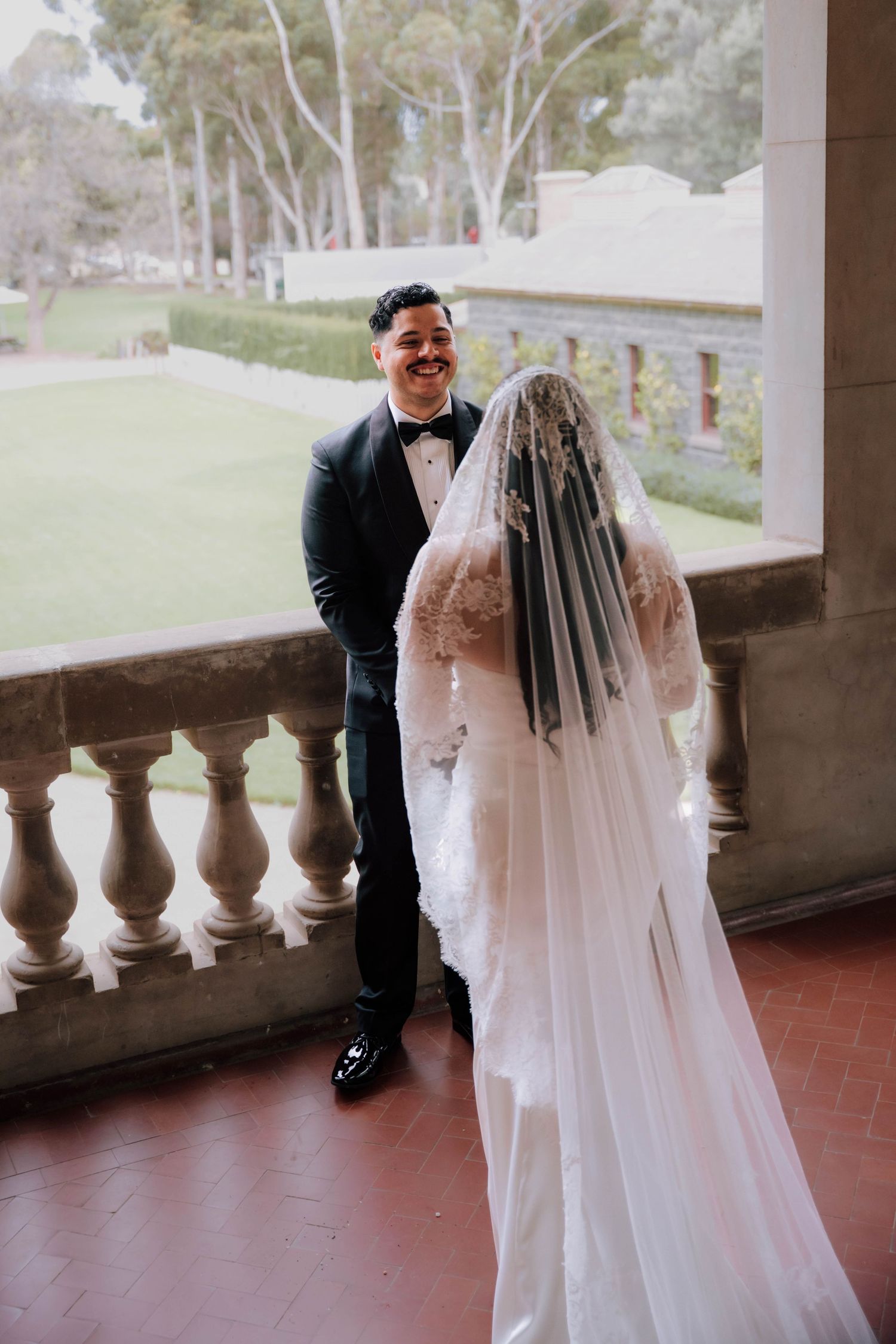 Groom smiling at bride in lace veil during outdoor wedding first look on balcony.