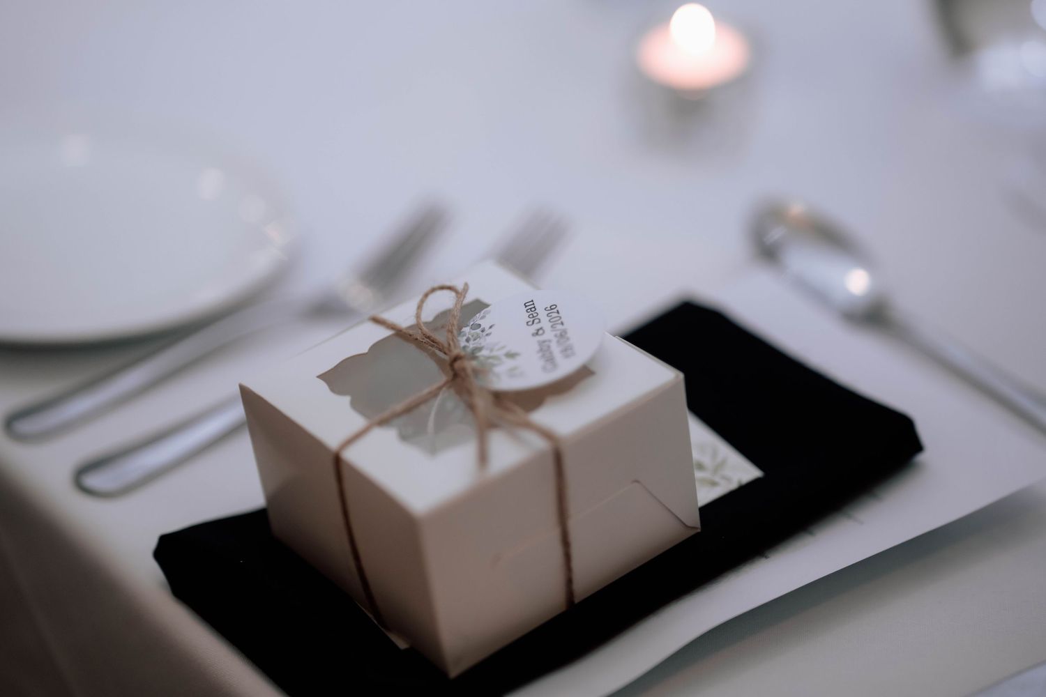 Elegant white wedding favor box tied with twine and a tag placed on a black napkin at a reception.
