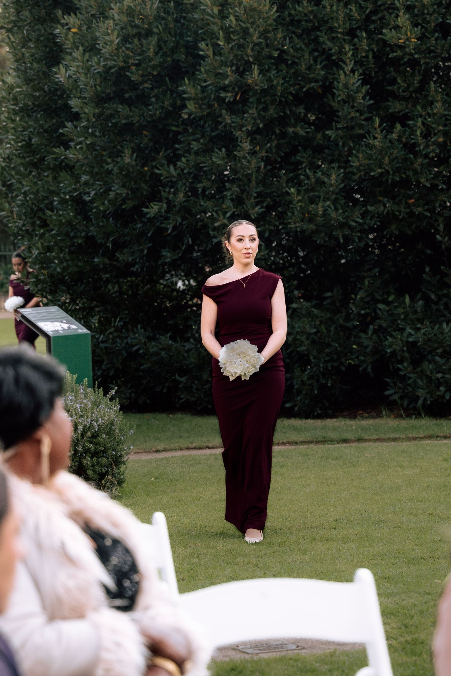 Bridesmaid in burgundy gown walking down outdoor wedding aisle carrying white bouquet.