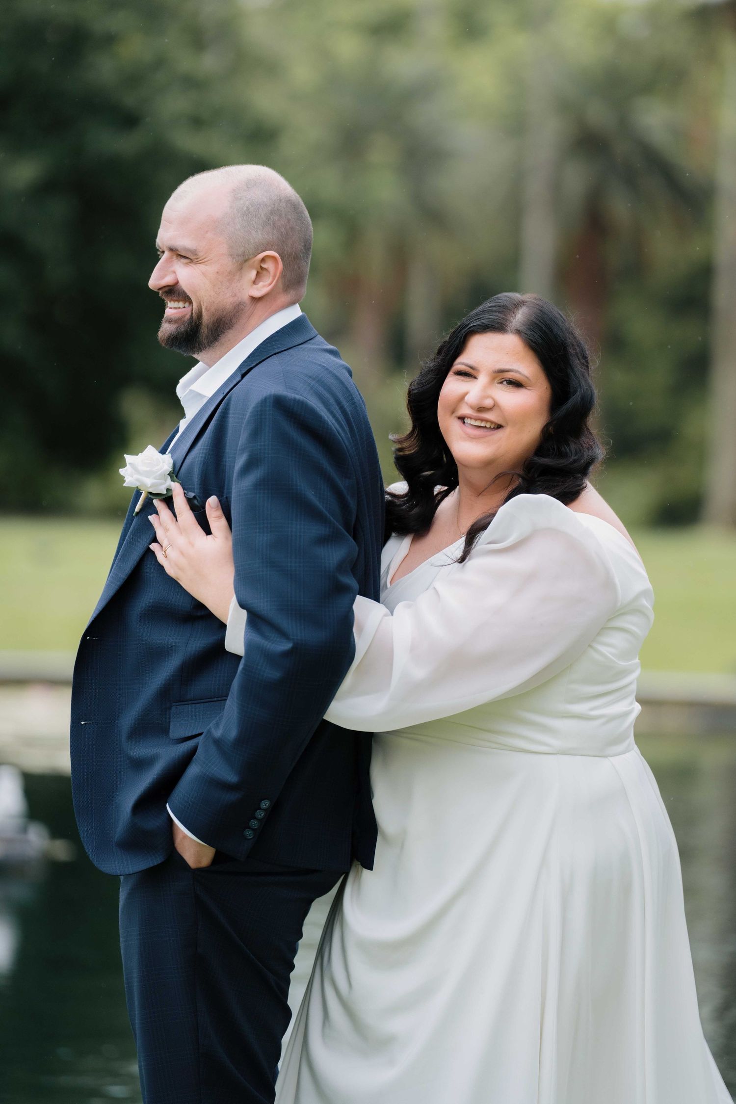Smiling bride embracing groom from behind near a tranquil pond in a green park.