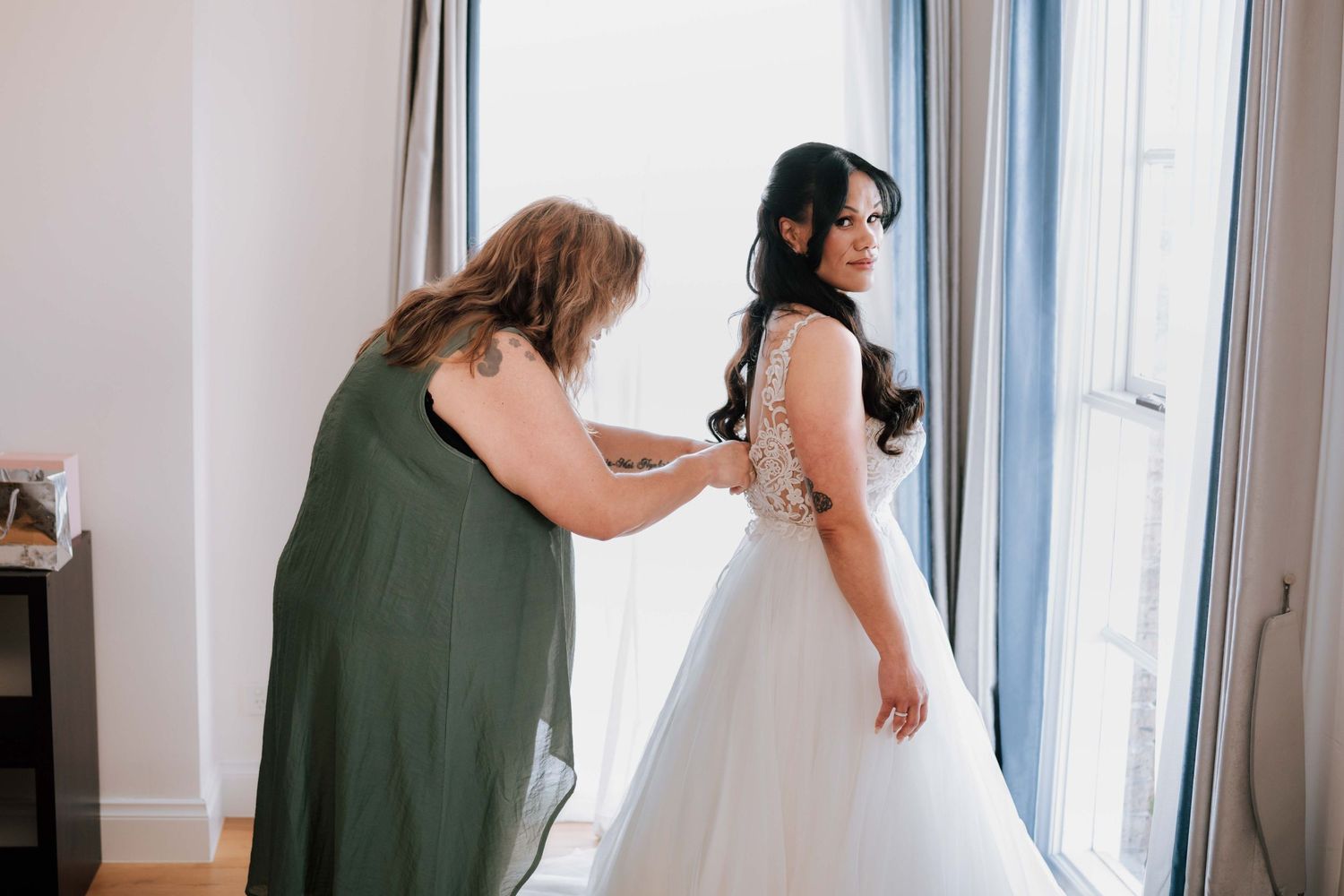 Mother helping bride fasten her white lace wedding gown near a bright window before the ceremony.