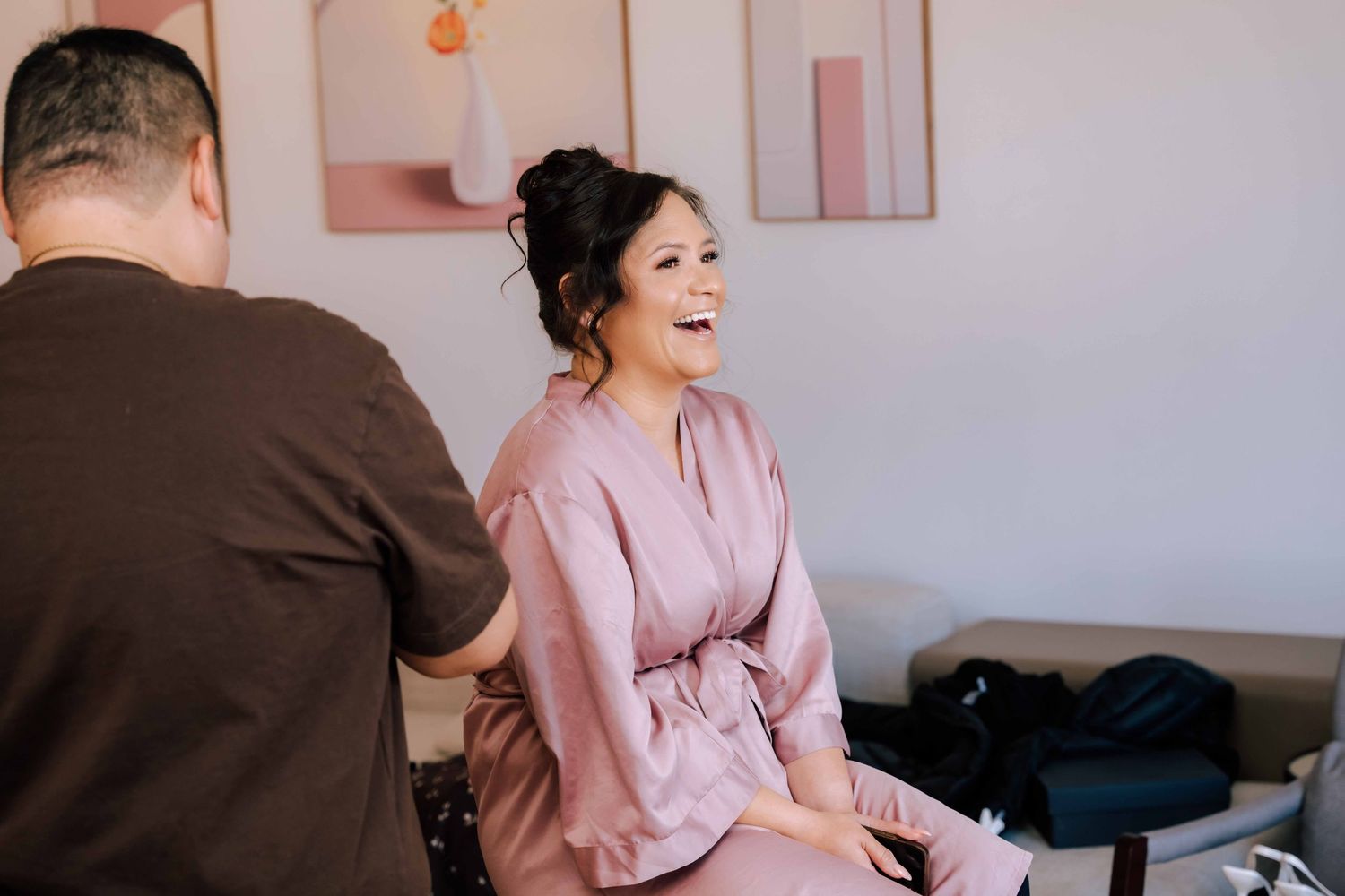 A woman in a pink satin robe laughs joyfully during bridal preparations in a modern bright room.