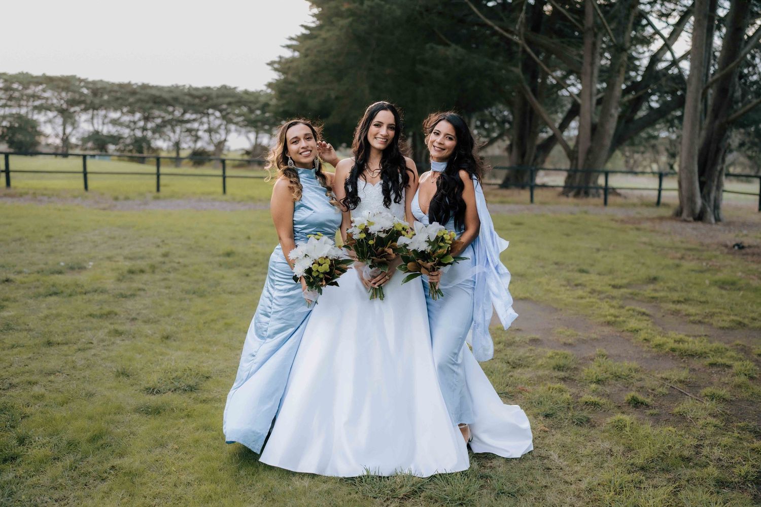 Bride and two bridesmaids in light blue dresses pose with bouquets on grass at outdoor wedding.