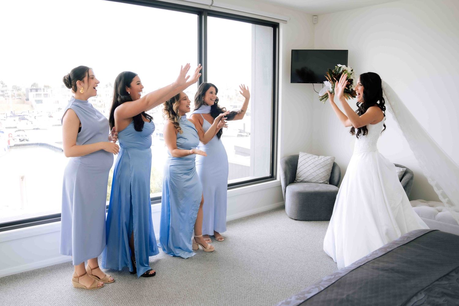 Bride in white gown reveals herself to bridesmaids in blue dresses in hotel room.