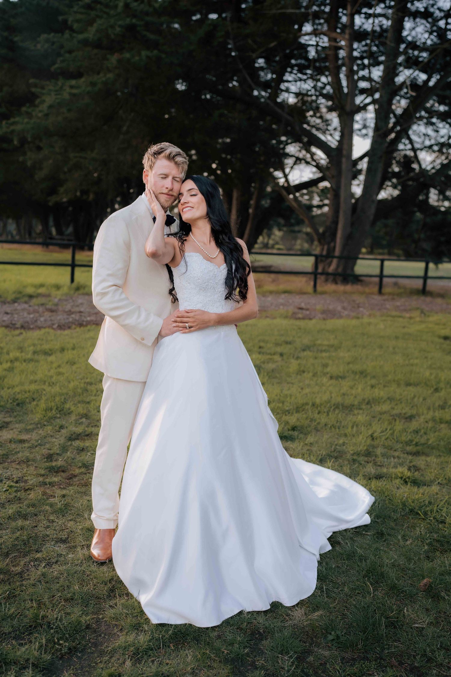 Bride and groom embrace outdoors on green grass at golden hour, groom in cream suit, bride in flowing white gown.