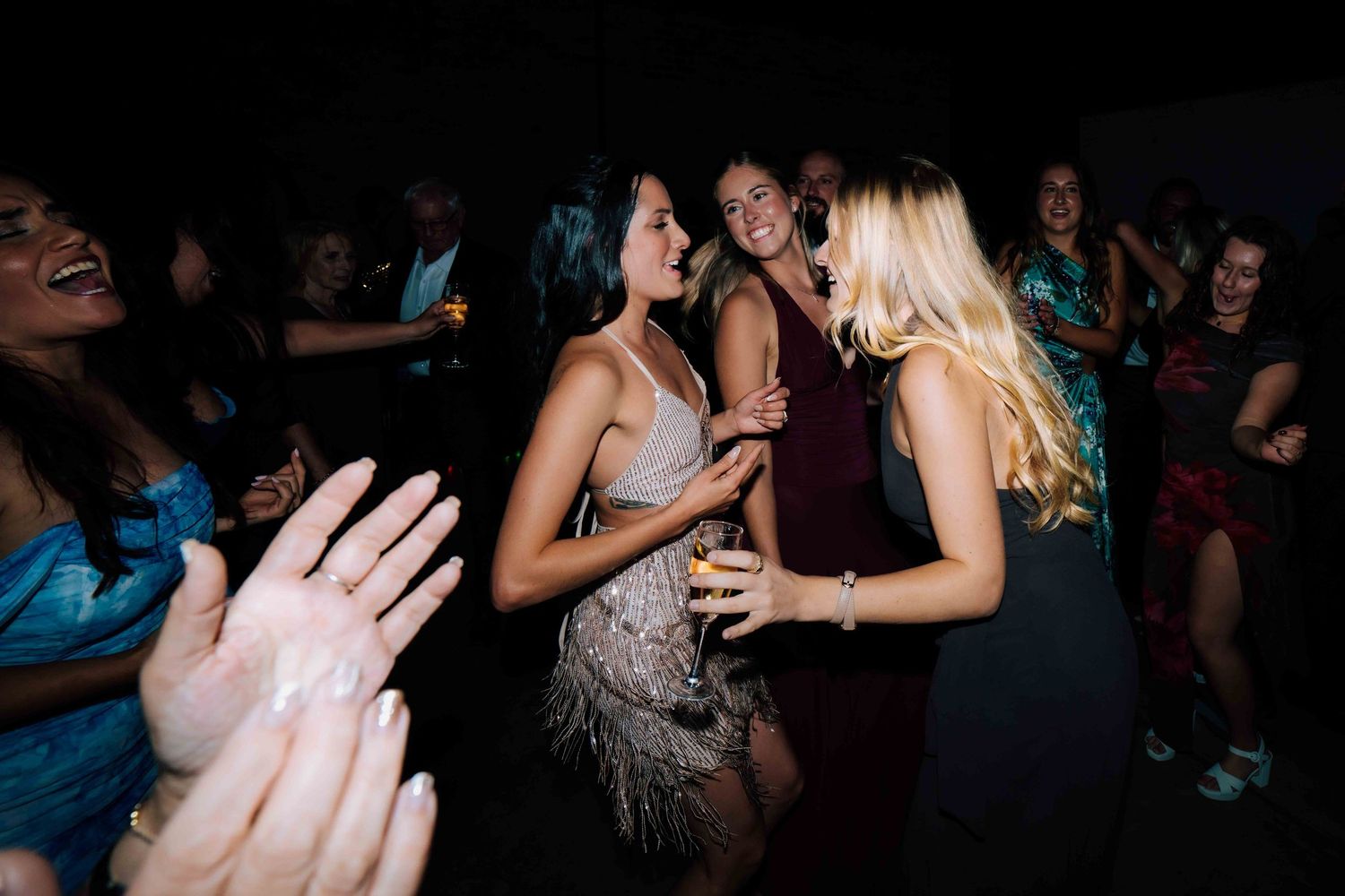 Women in elegant dresses laugh and dance together at a lively wedding reception party.
