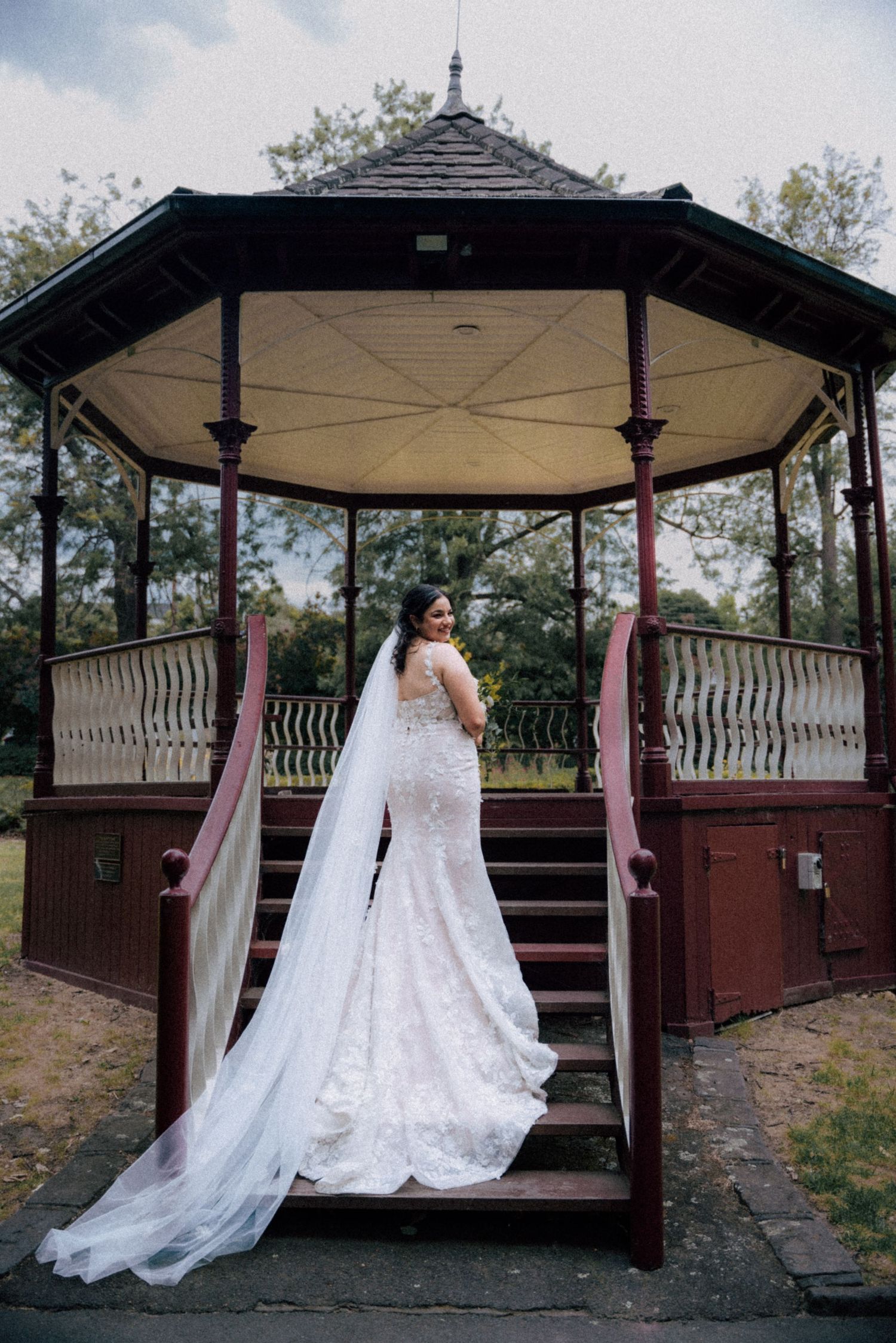 Bride in lace mermaid gown with long veil stands on steps of vintage red gazebo, back to camera.