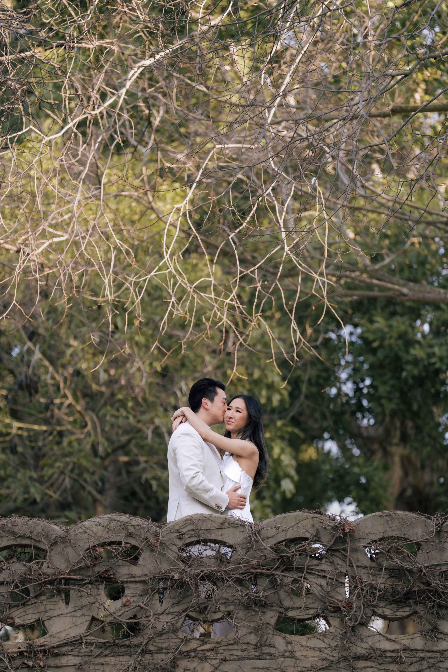 Asian couple in white wedding attire share a kiss beside a stone wall surrounded by lush garden trees.