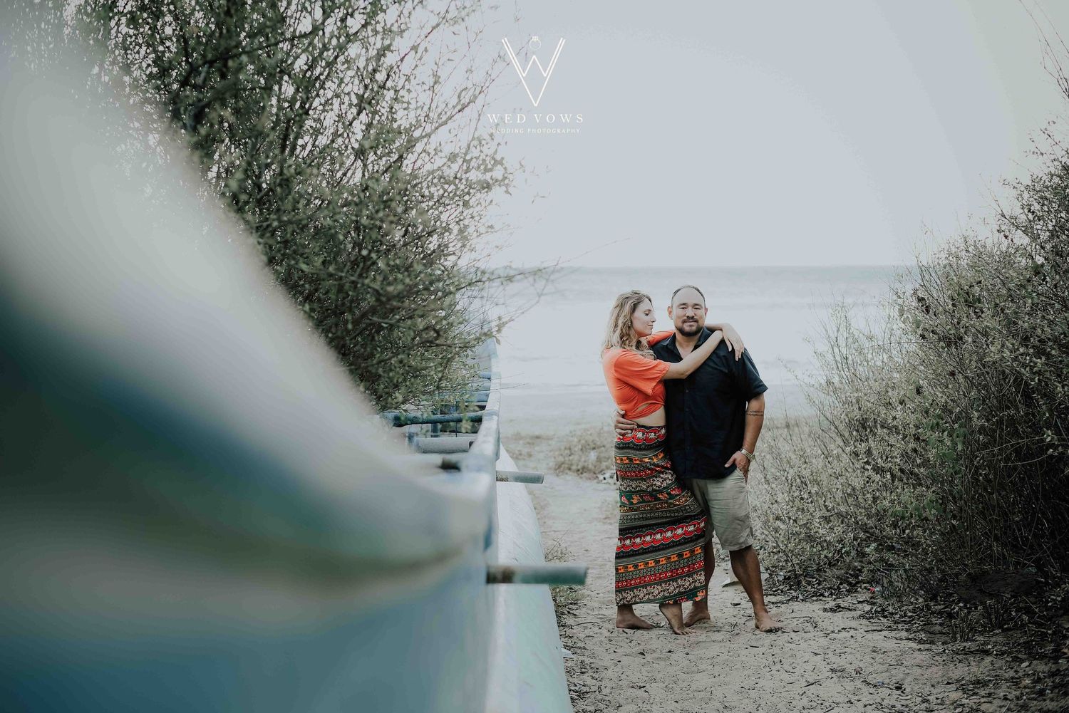Couple sharing a tender moment on a sandy path surrounded by lush greenery near the ocean.