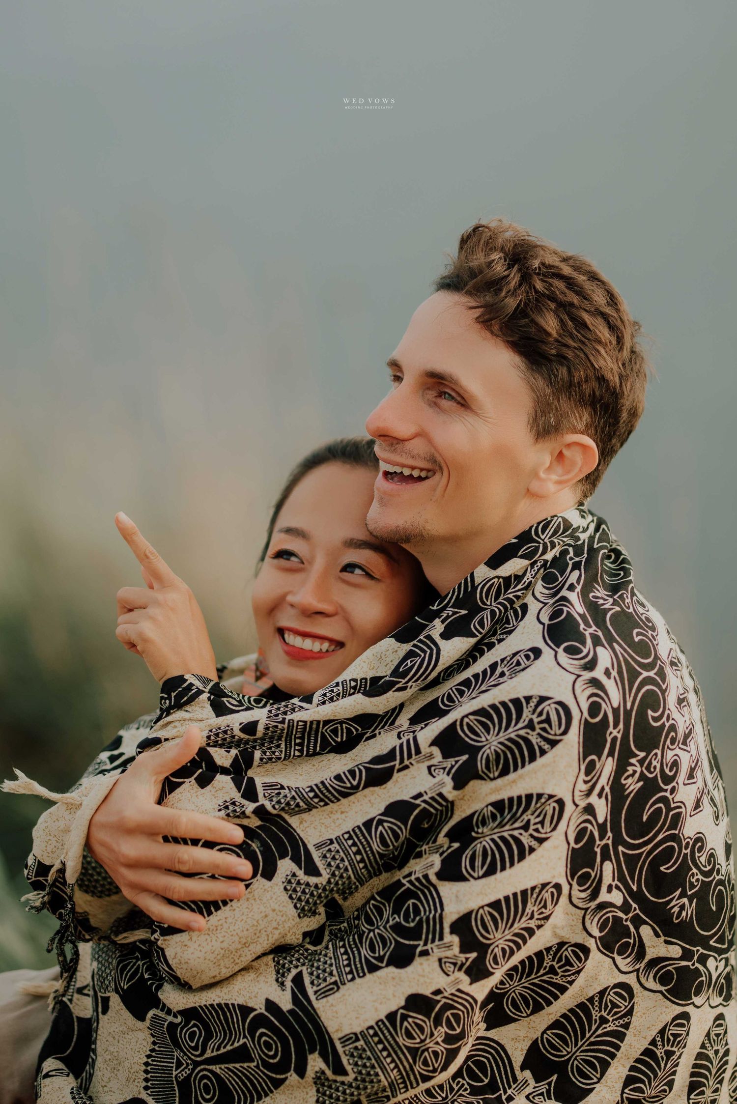 Couple wrapped in black and white patterned blanket smiling and pointing outdoors.