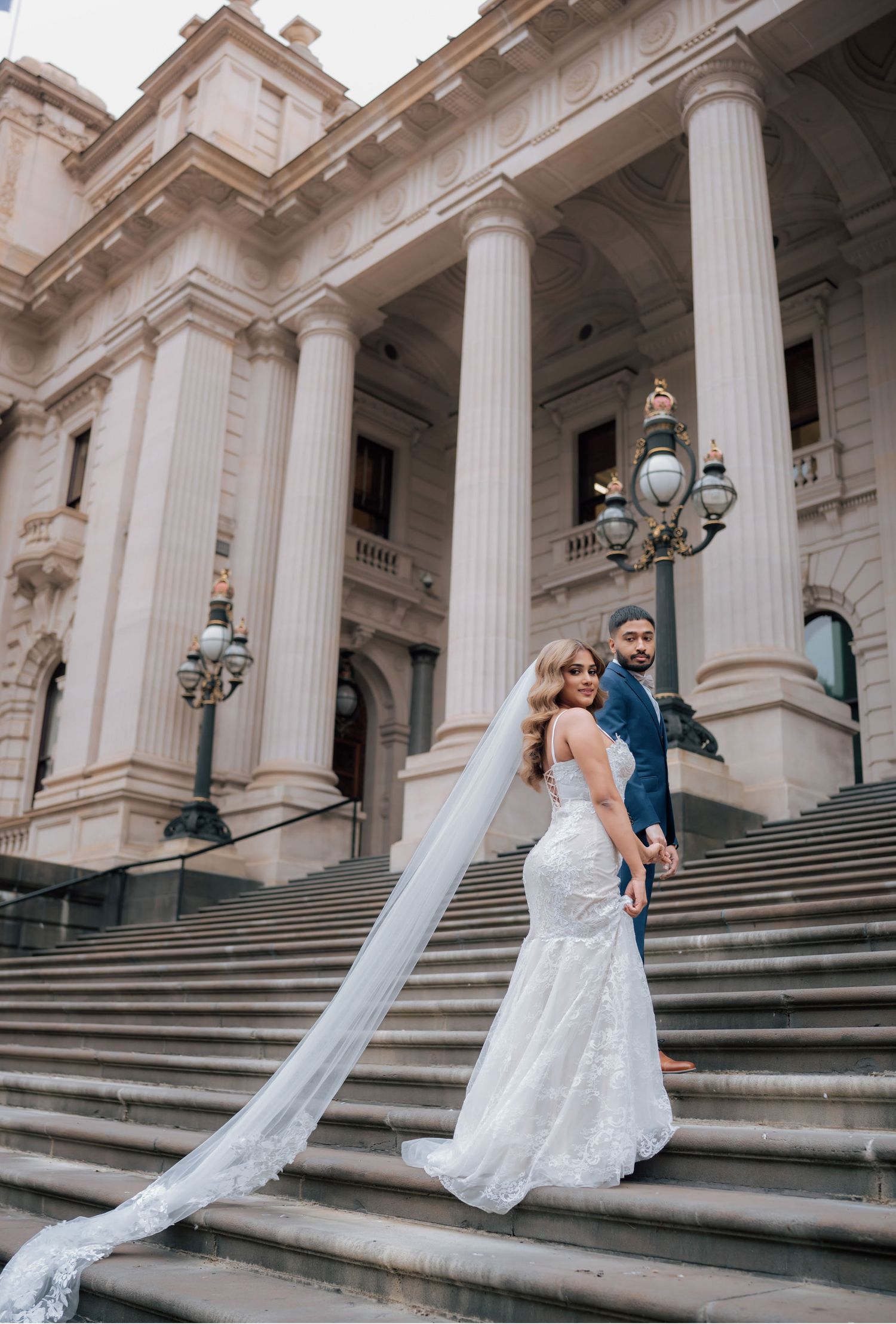 Bride in lace mermaid gown with long veil and groom in navy suit on grand building steps.