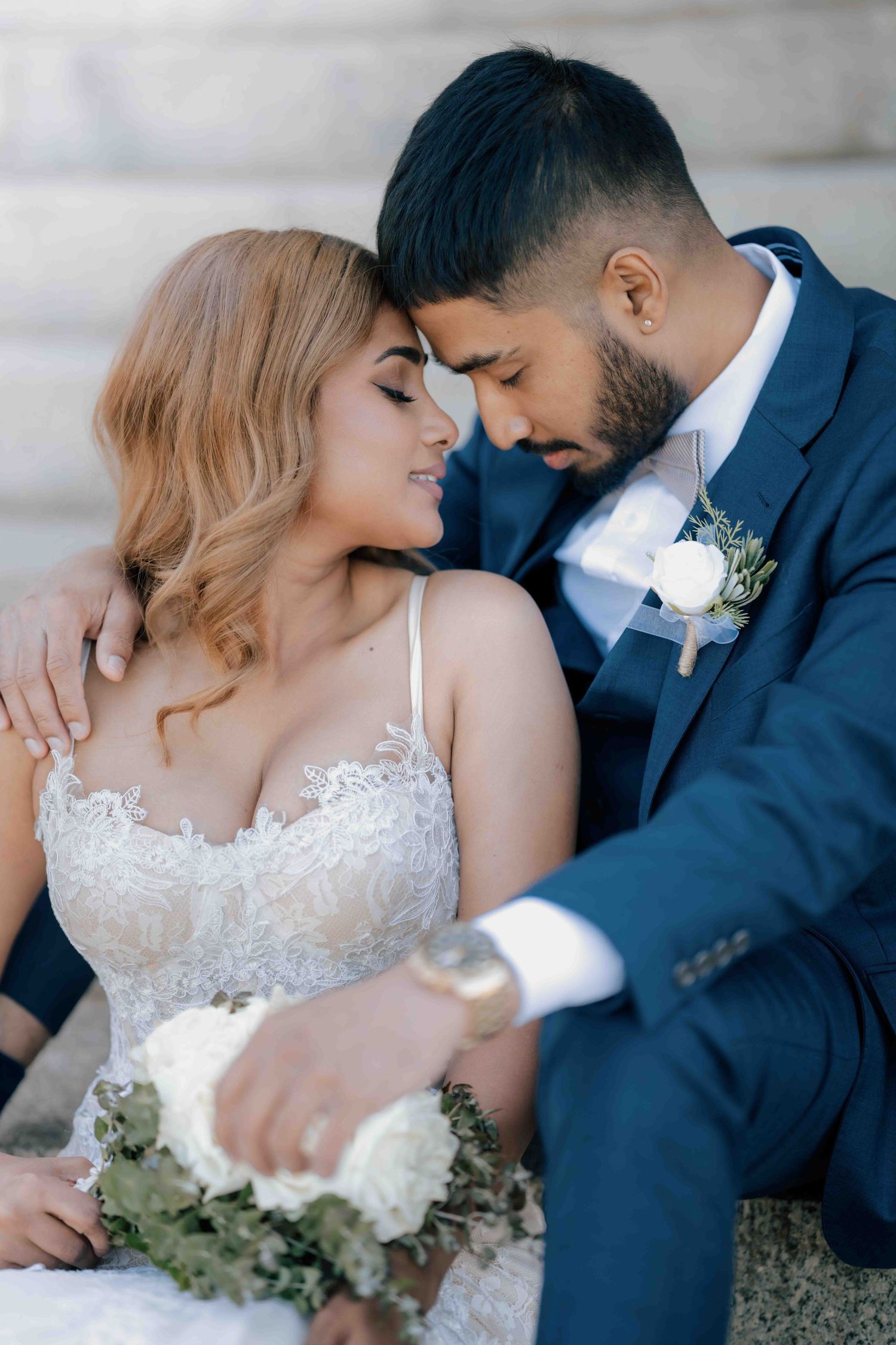 Wedding couple sitting close together, bride holding white bouquet, groom in navy suit with boutonniere.