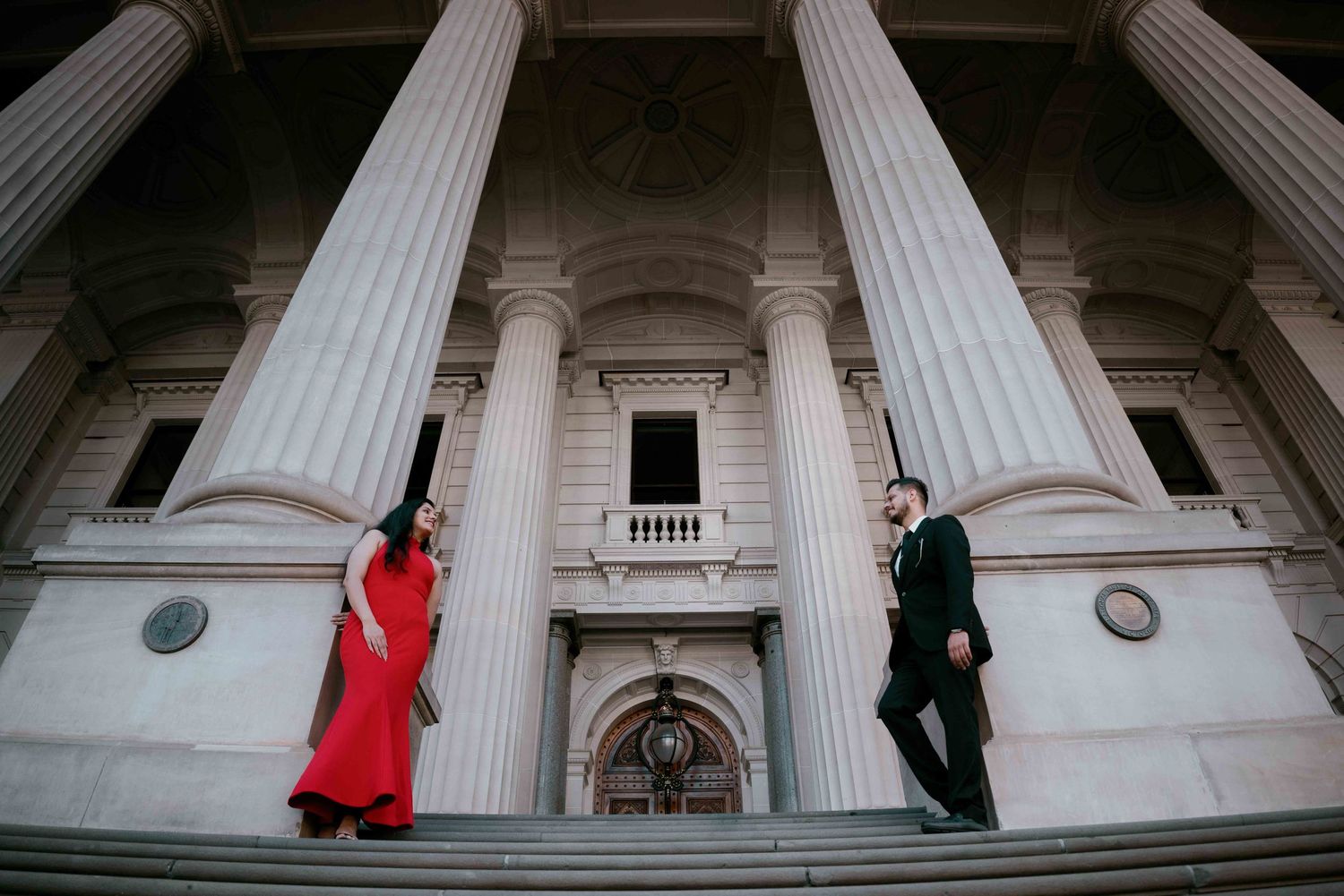 Elegant couple in red dress and black suit posing on steps of grand neoclassical building with tall columns.