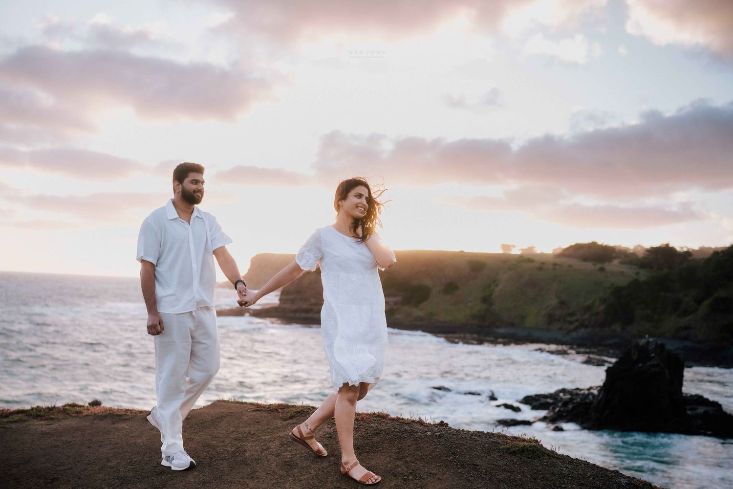 Couple in matching white outfits walking hand in hand along rocky coastal cliffs at sunset.