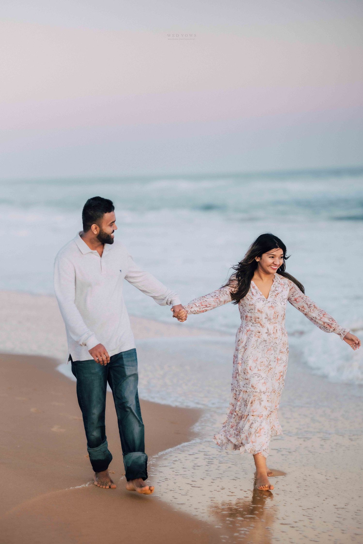 Couple walking hand in hand along the shoreline with ocean waves in the background at dusk.