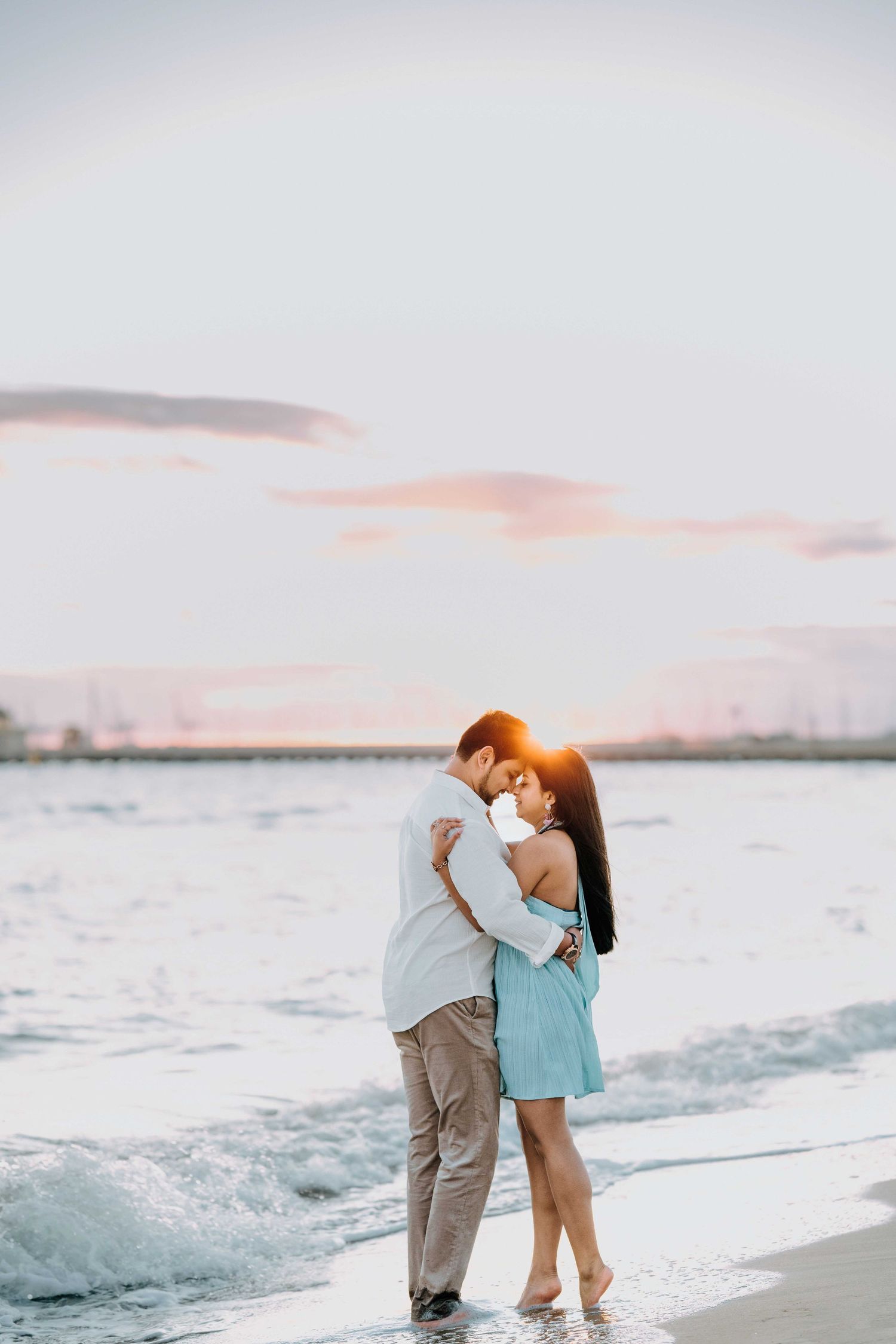 Couple embracing romantically on beach shore at golden sunset, woman in blue dress, man in white shirt.