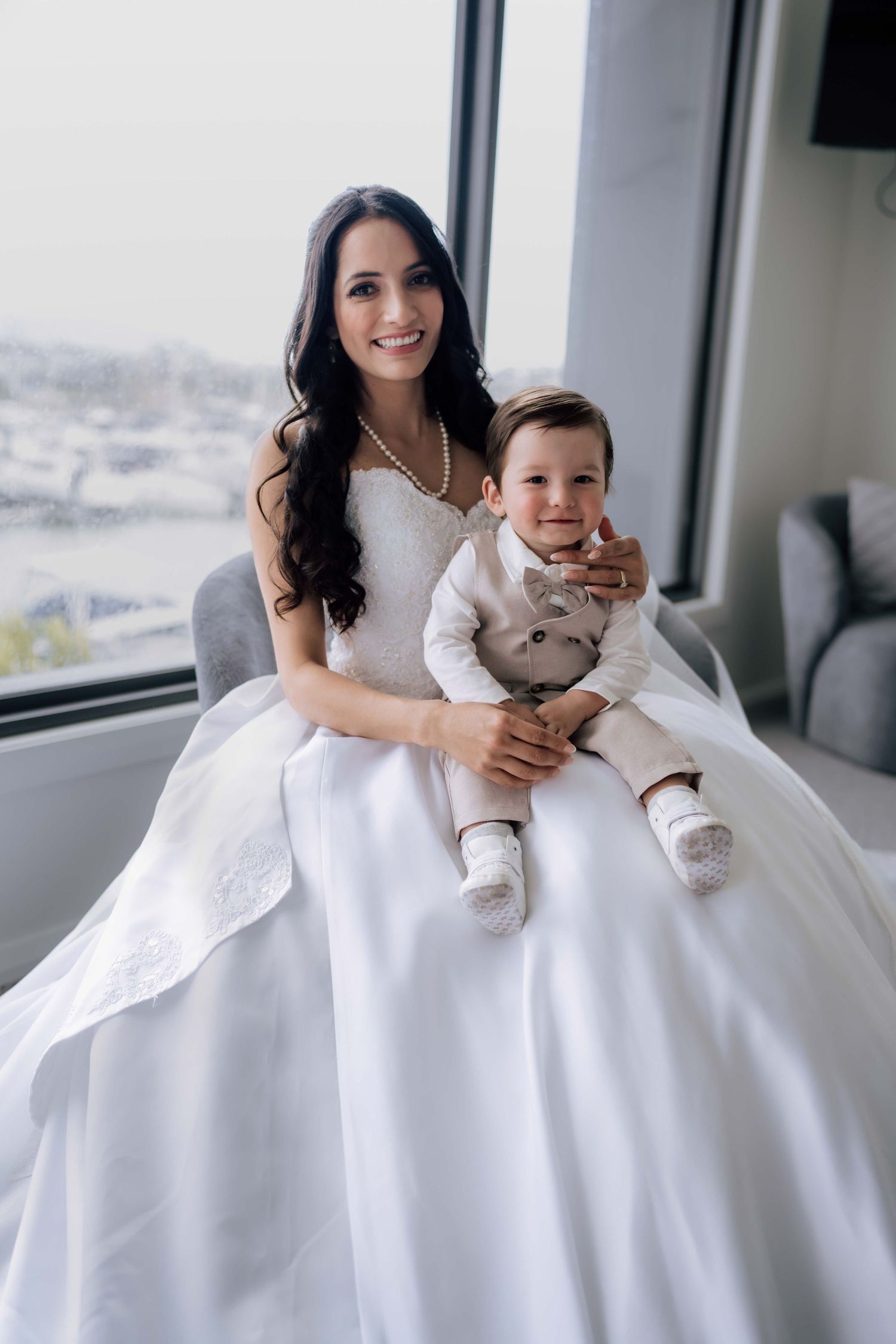 Radiant bride in white ball gown sitting with a smiling baby boy dressed in a suit.