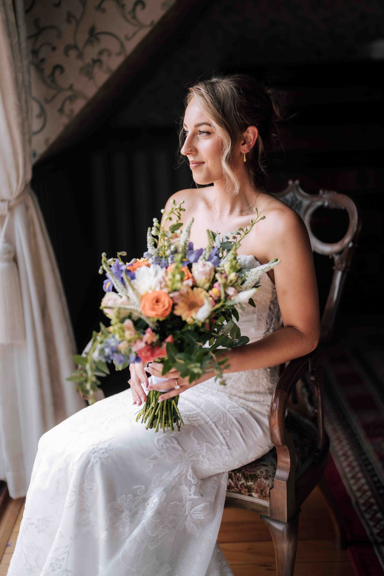 Elegant bride in lace wedding gown holding colorful bridal bouquet while seated in ornate chair.