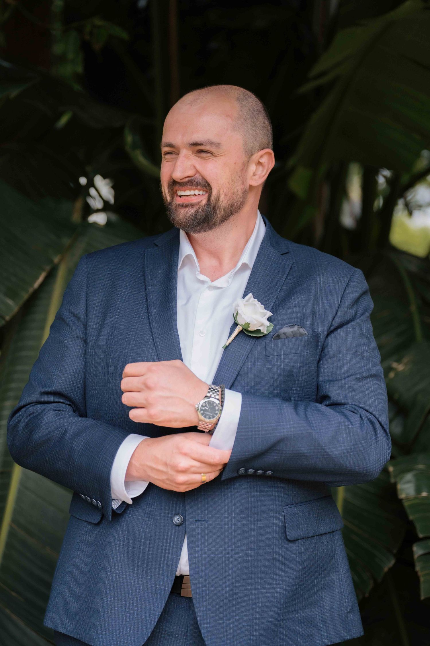 Cheerful bald groom in blue suit adjusting his cufflinks with a boutonniere against tropical green foliage.