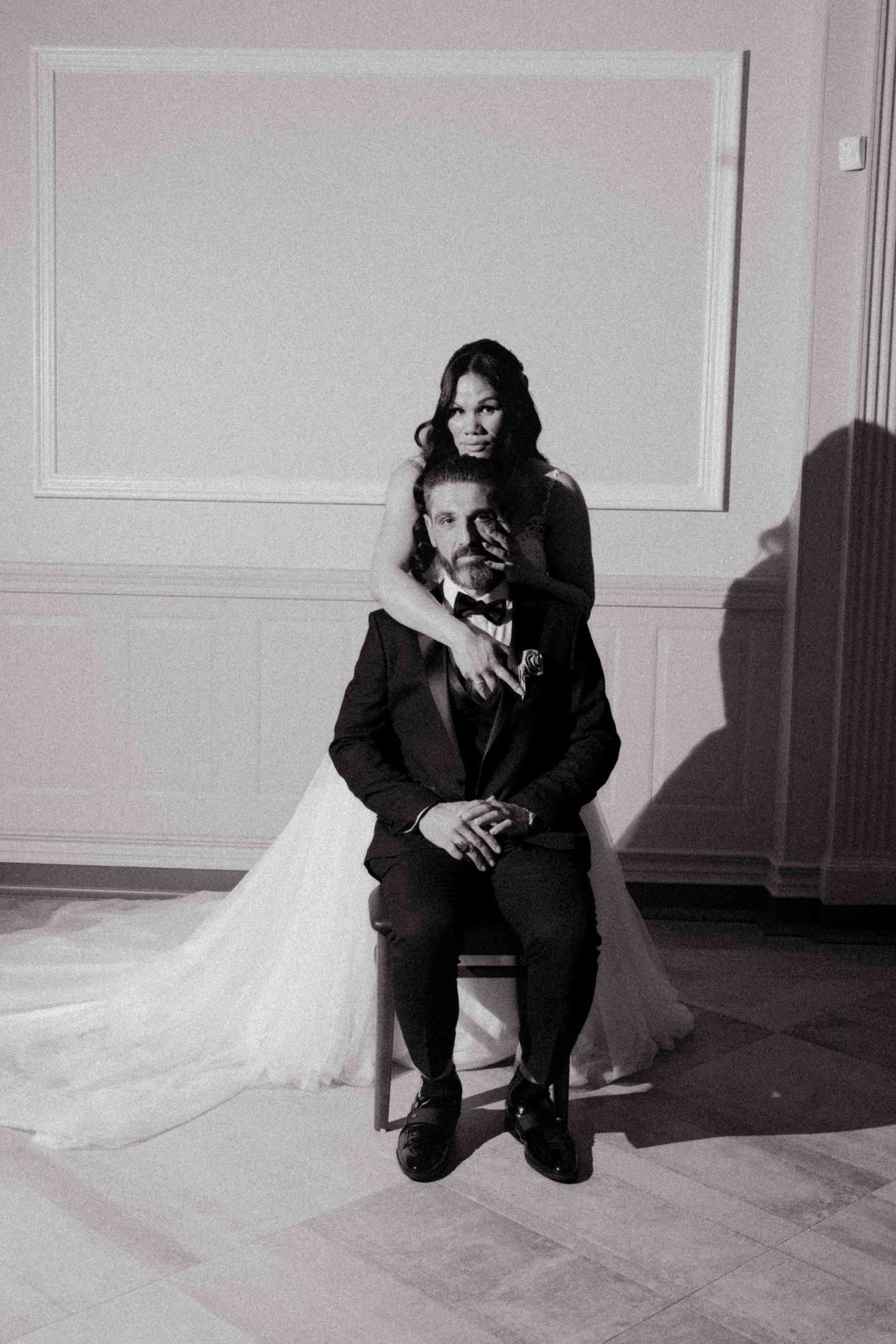 Black and white photo of bride and groom posing elegantly, groom seated while bride stands behind him in a ballroom.