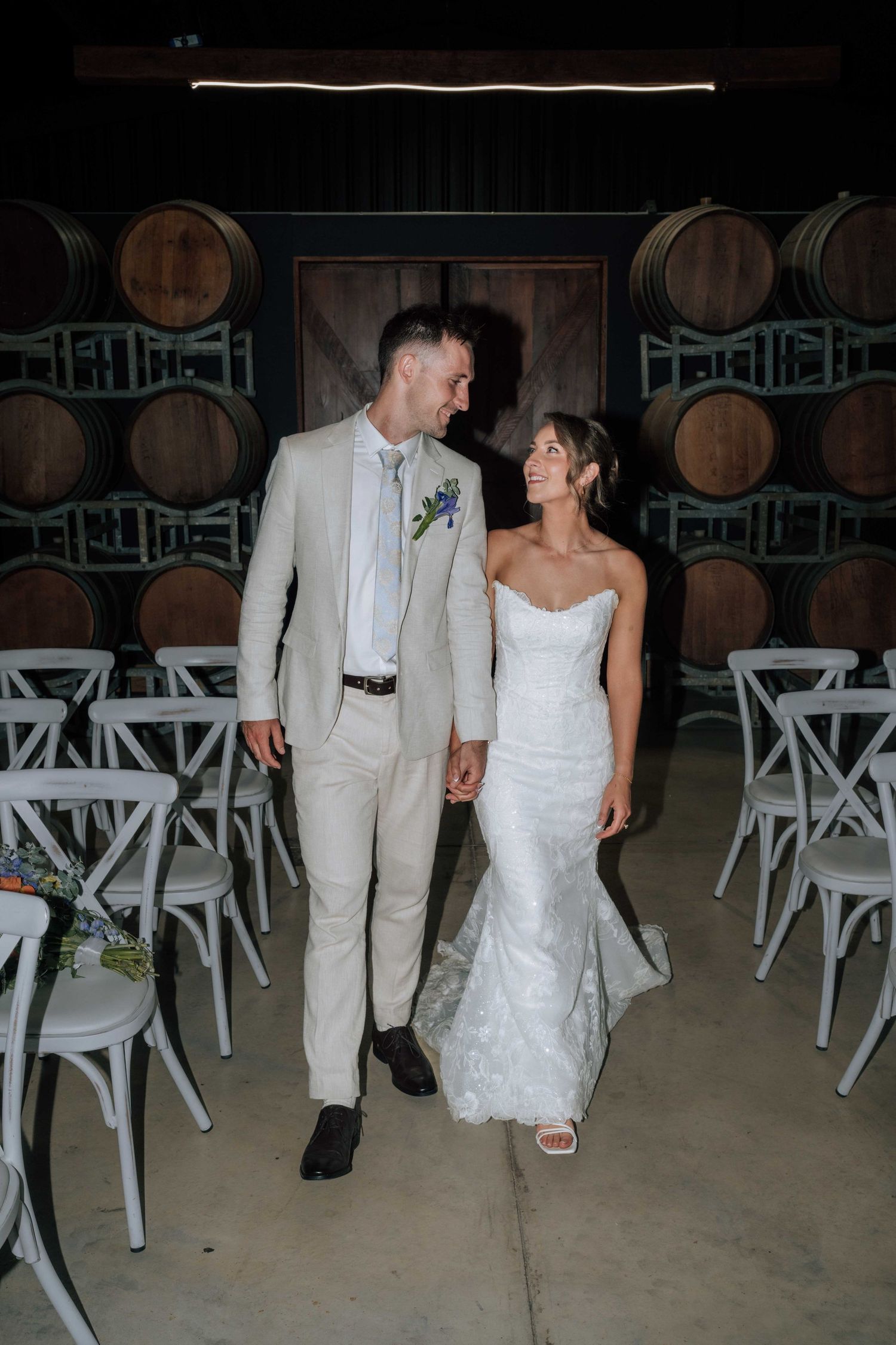 Bride in lace mermaid gown and groom in tan suit walk hand in hand through winery barrel room.